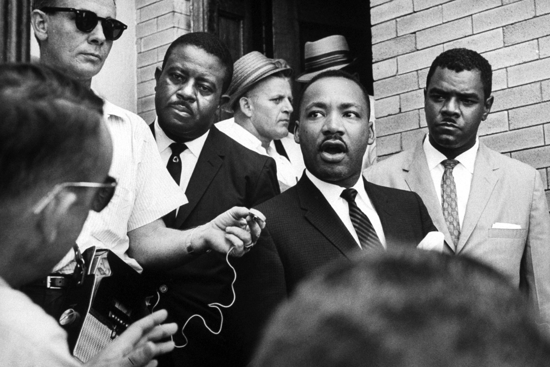 A man speaks to reporters holding microphones outside a brick building, surrounded by other men in suits, conveying a serious atmosphere.