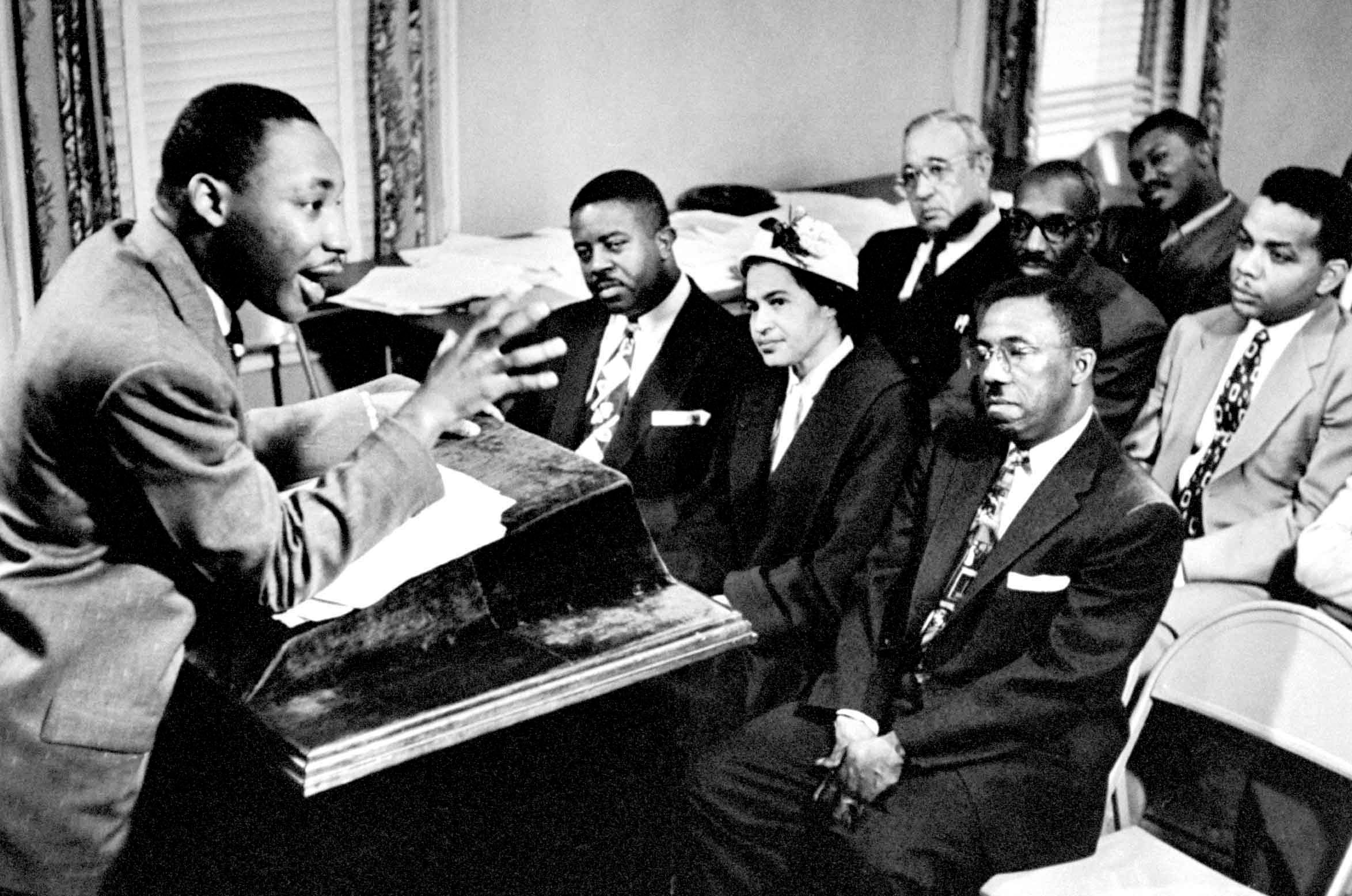 Man speaks passionately at podium; seated audience listens intently in a small, furnished room with visible papers and chairs.