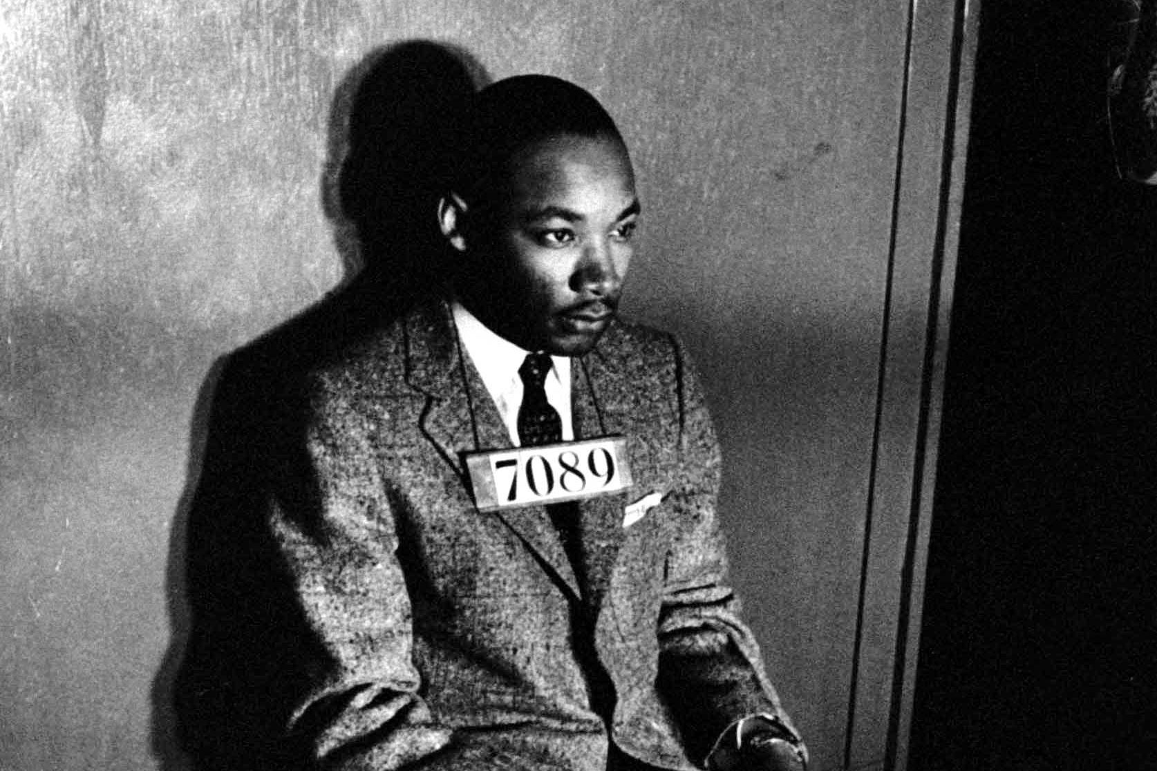 Man sits wearing suit with "7089" tag, gazing thoughtfully, in a dimly lit room.