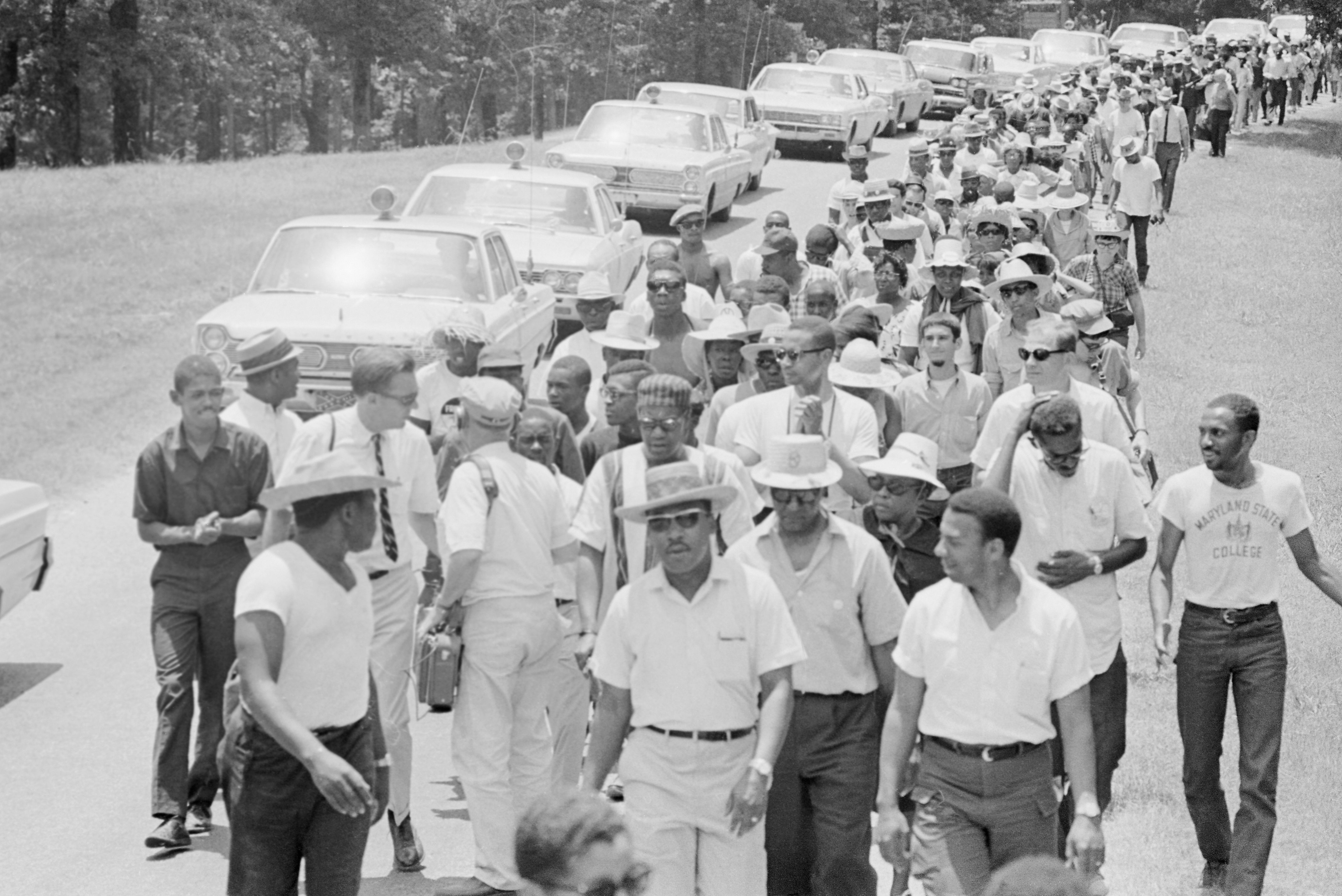 A large group of people walks along a road, surrounded by parked cars, with trees in the background.