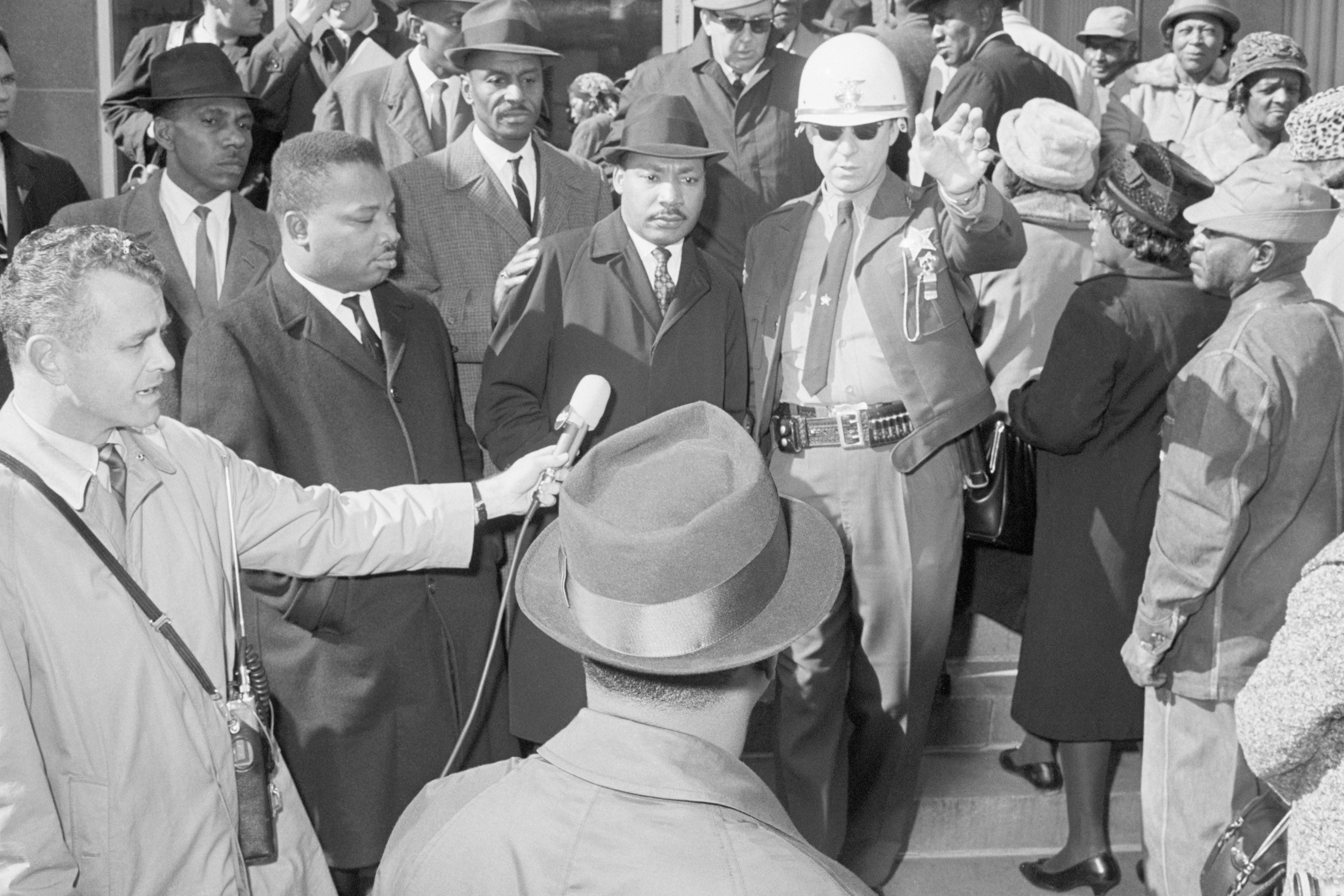 Police officer directs a crowd while a man speaks into a microphone, surrounded by people in coats and hats on outdoor steps.