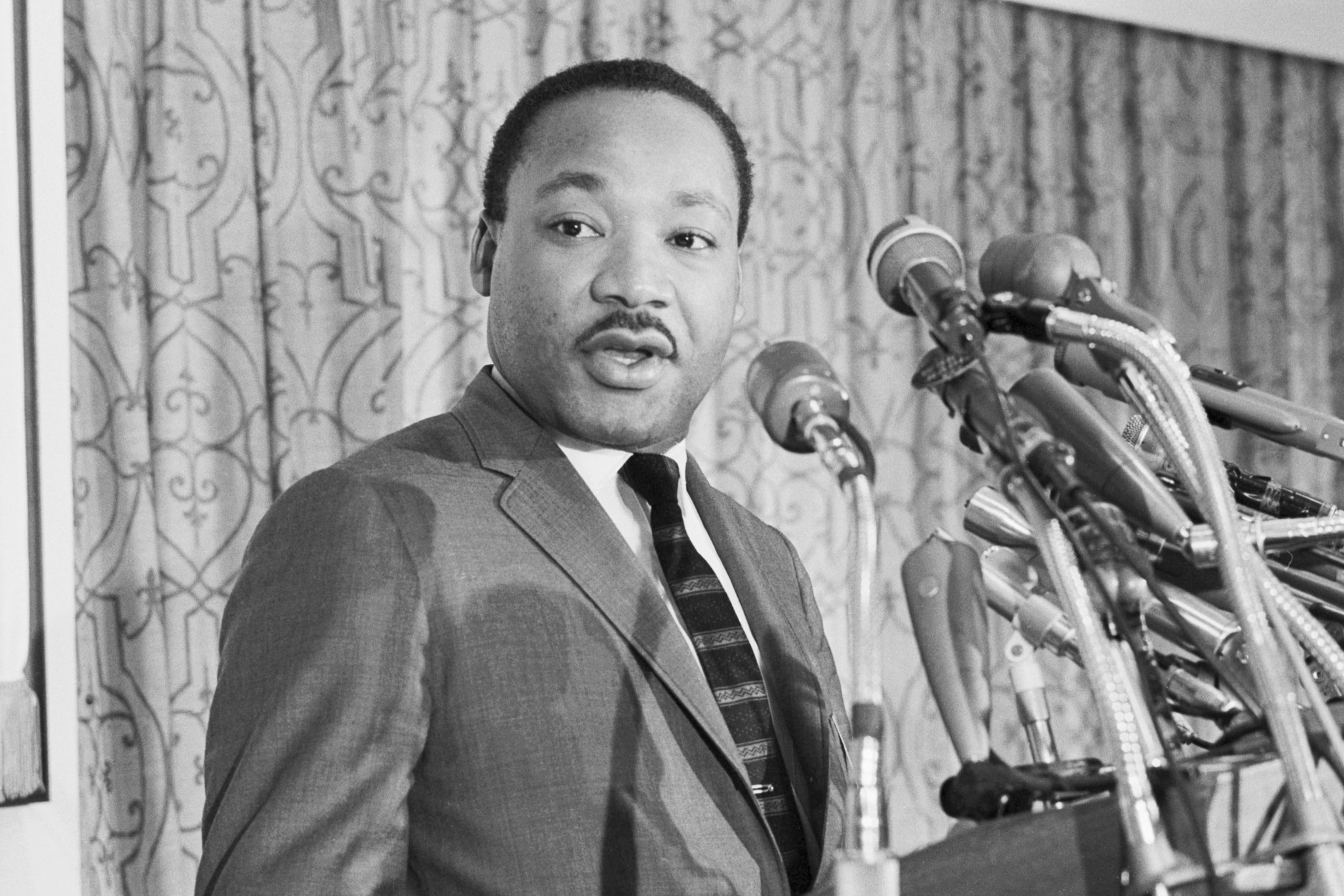 A man speaks into multiple microphones, wearing a suit and tie, with a patterned curtain in the background.