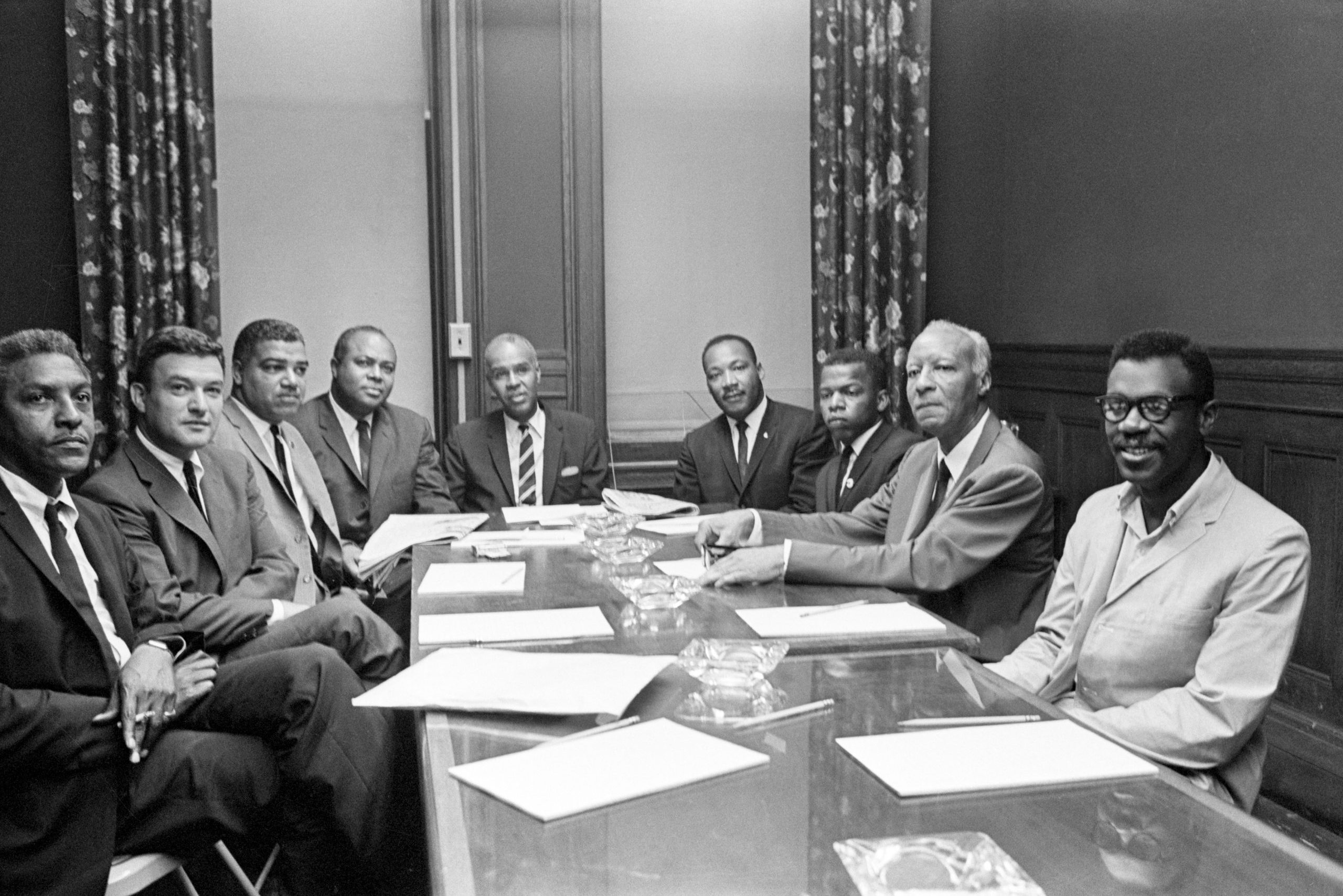 Eight men sit at a table, engaging in a meeting in a room with curtains and wood-paneled walls. Papers are on the table.