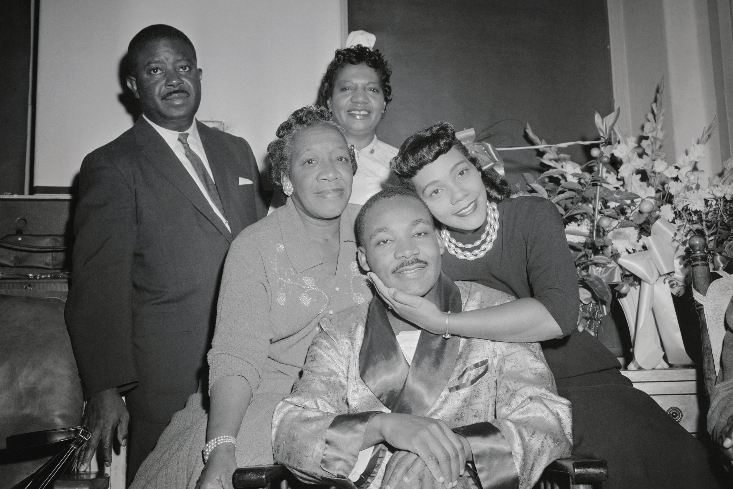 A man in a patterned robe sits surrounded by smiling people, with flowers in the background. A woman lovingly embraces him.