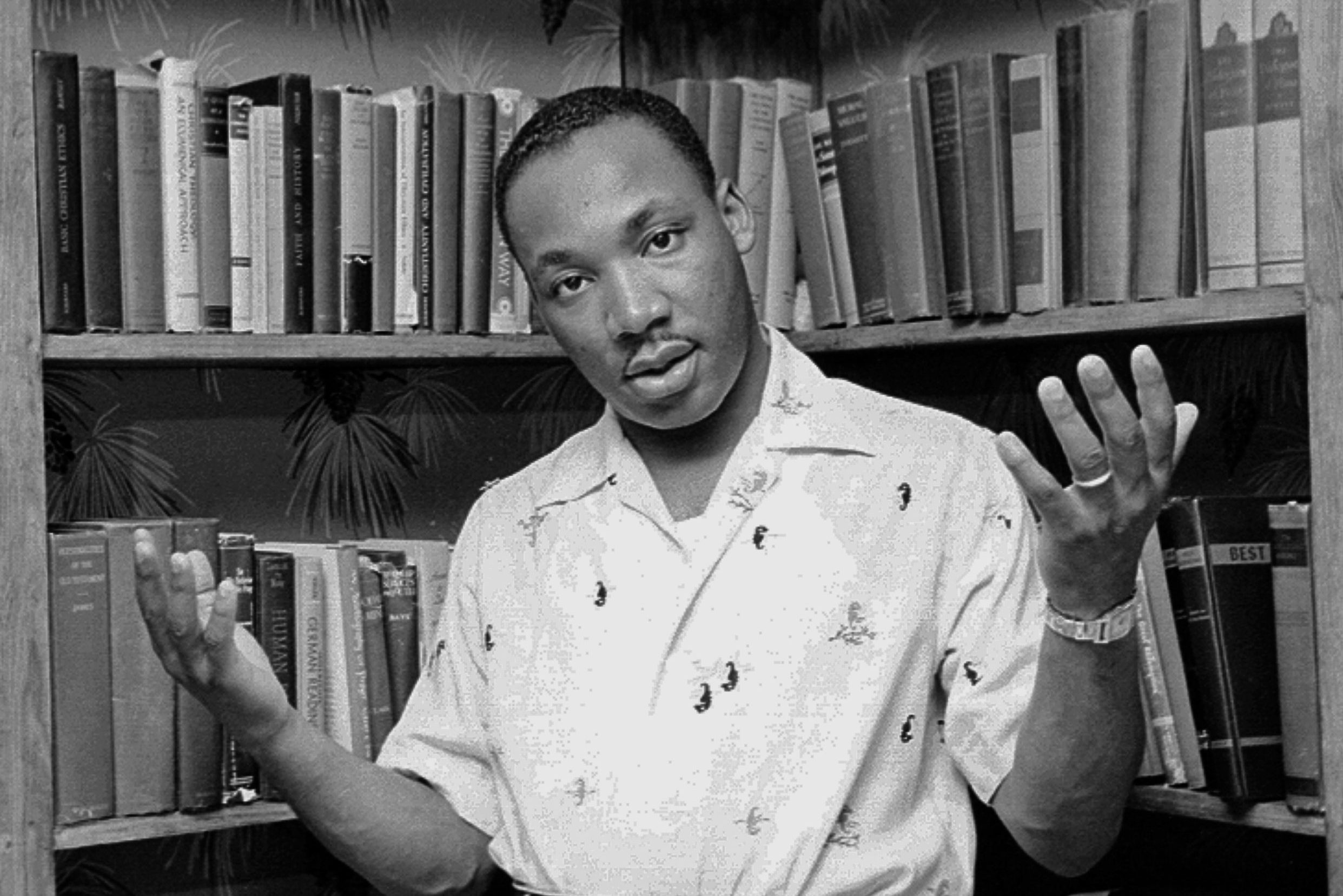 Person gestures with open hands in front of a bookshelf filled with various books. They wear a patterned shirt in a casually posed setting.