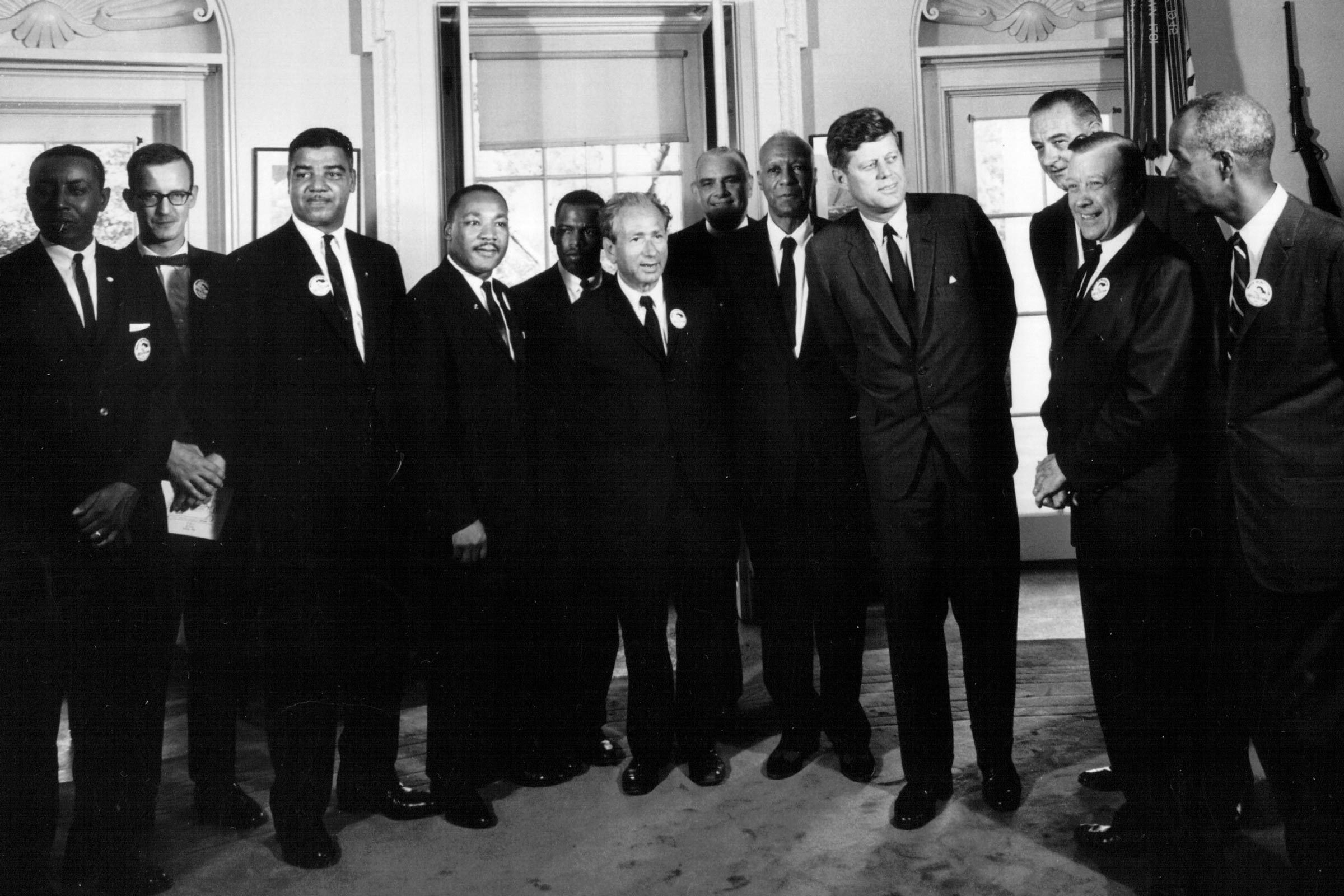 A group of men in suits stand together, posing for a photo in an ornate room with a window in the background.