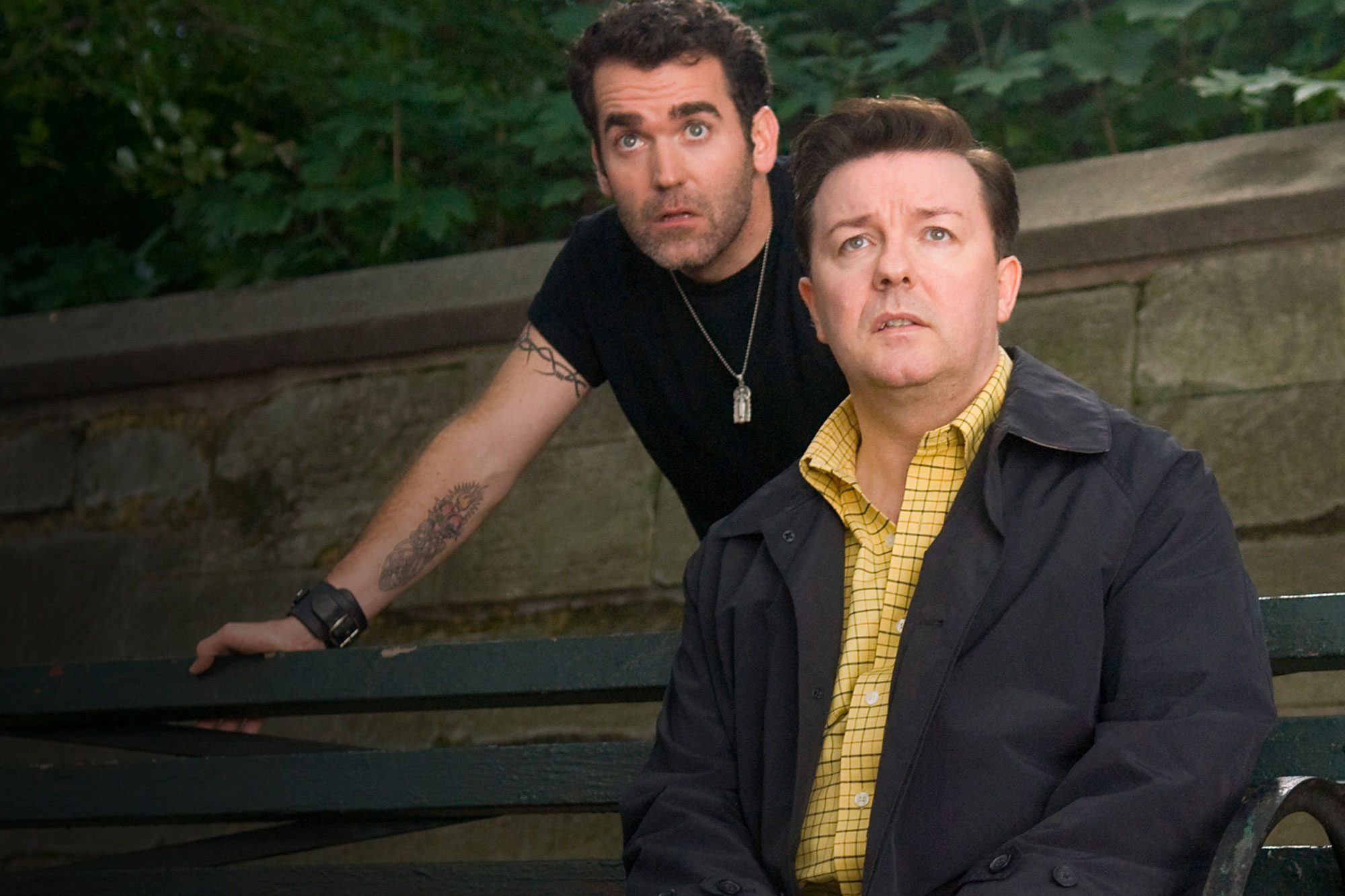 Two men look upward, appearing surprised, in a park setting with a stone wall and greenery in the background.