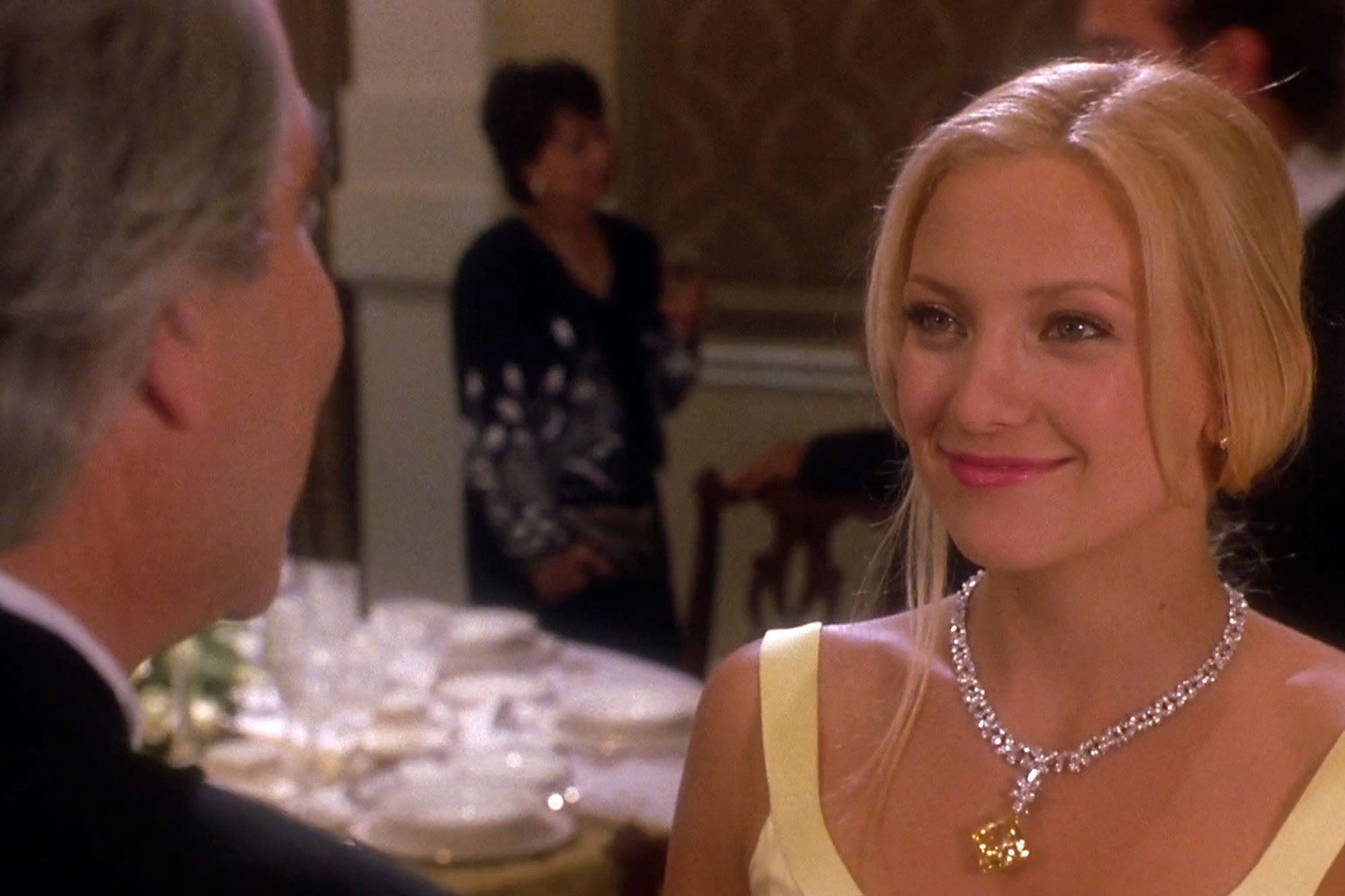 A woman smiles at a man, inside a formal dining room with set tables and guests in the background.