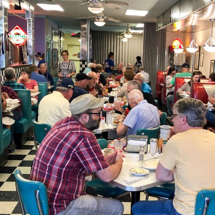 Diners sit and eat at a busy 1950s-style diner with teal booths, checkered floor, and retro decor.
