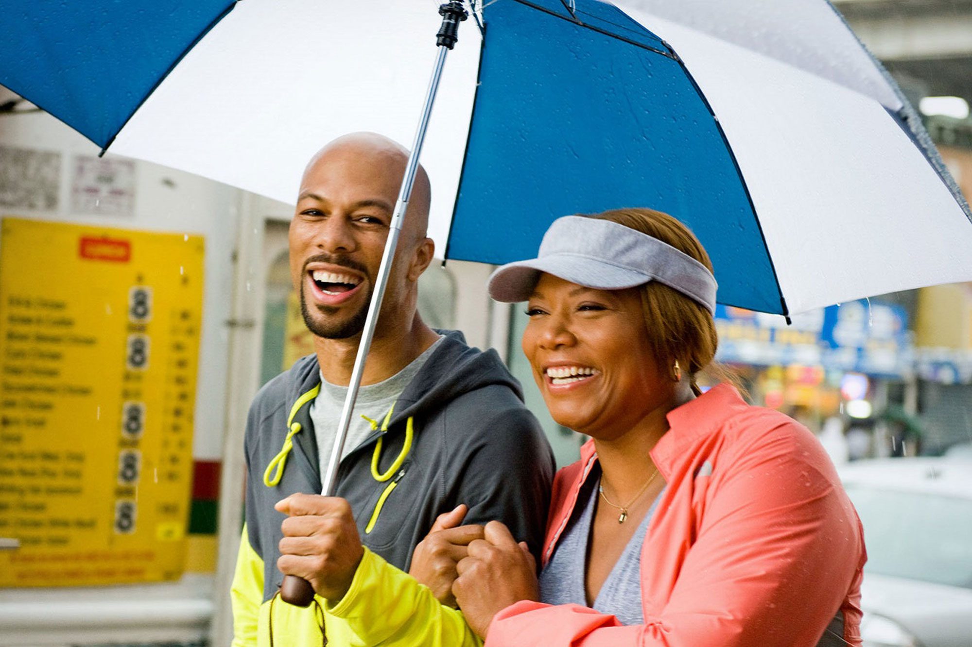 Two people walk under an umbrella, smiling and laughing, on a rainy street with blurred signage in the background.
