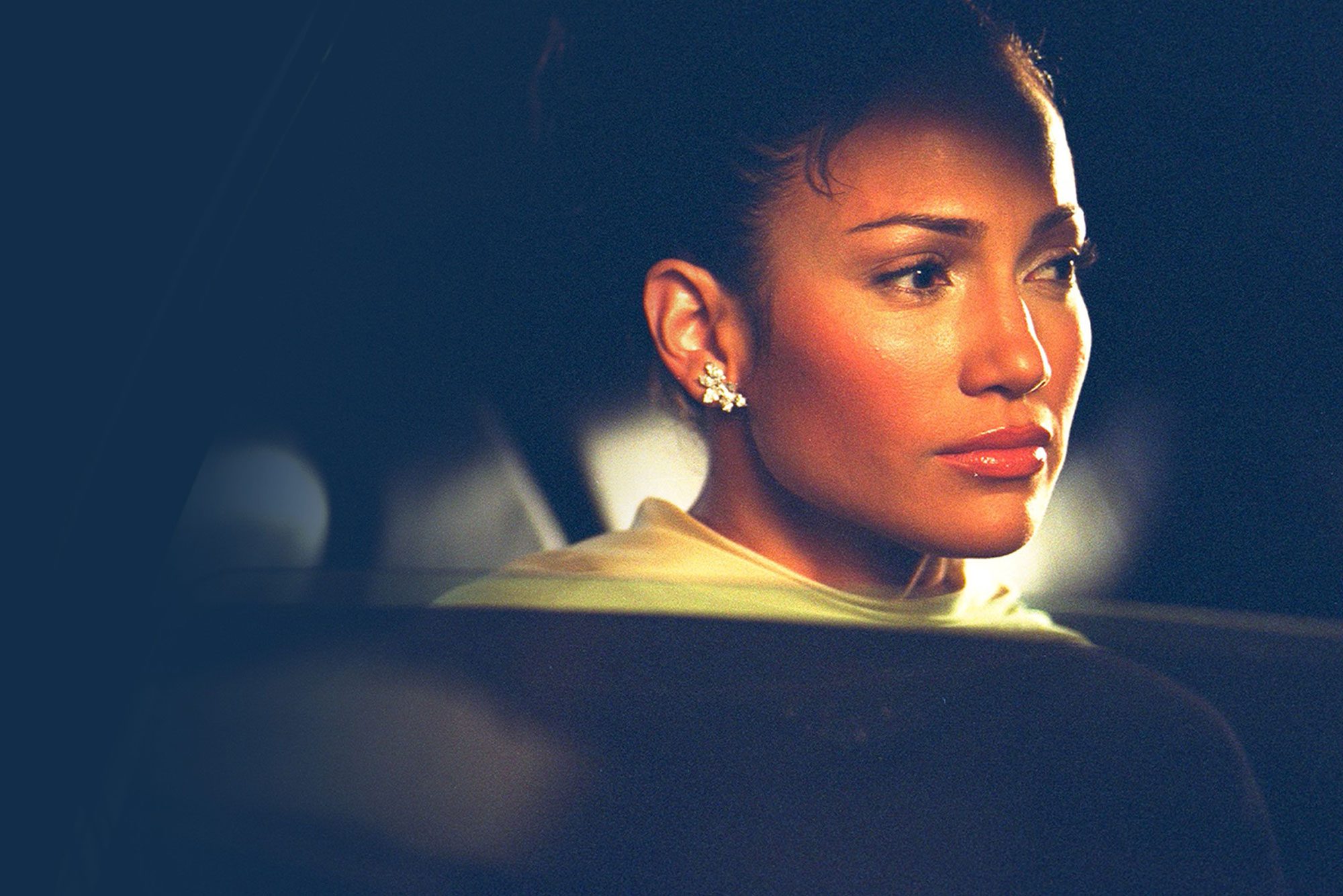 A woman looks pensively out a car window at night, softly lit, with earrings reflecting light.