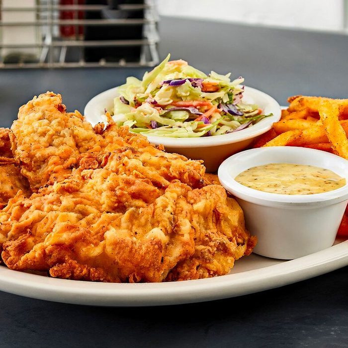 Fried chicken sits on a plate with coleslaw, seasoned fries, and a small bowl of dipping sauce on a dark tabletop.