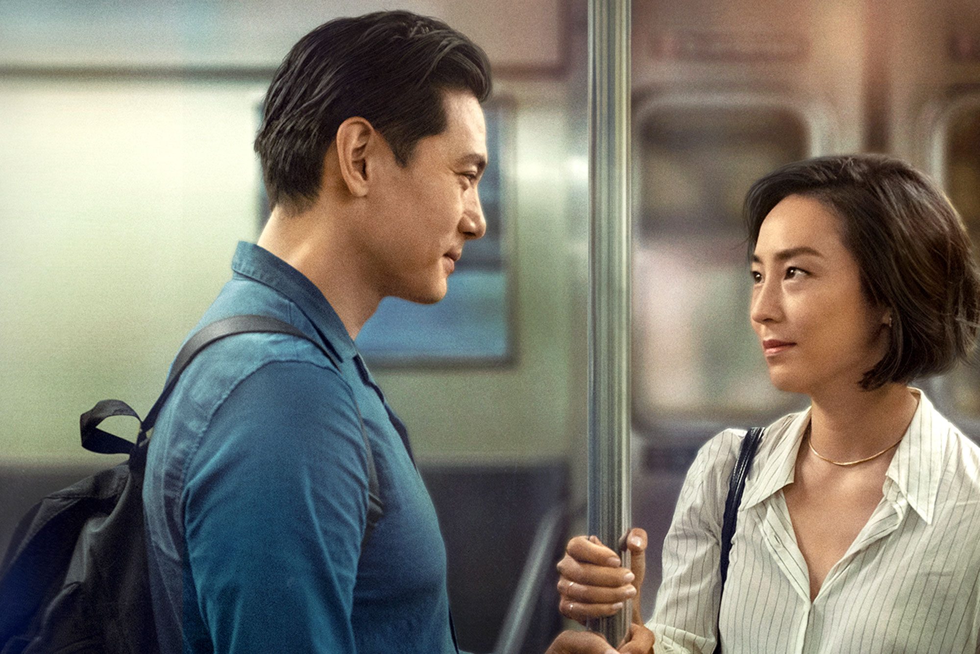 Two people gaze at each other, standing in a subway train, one holding a pole.