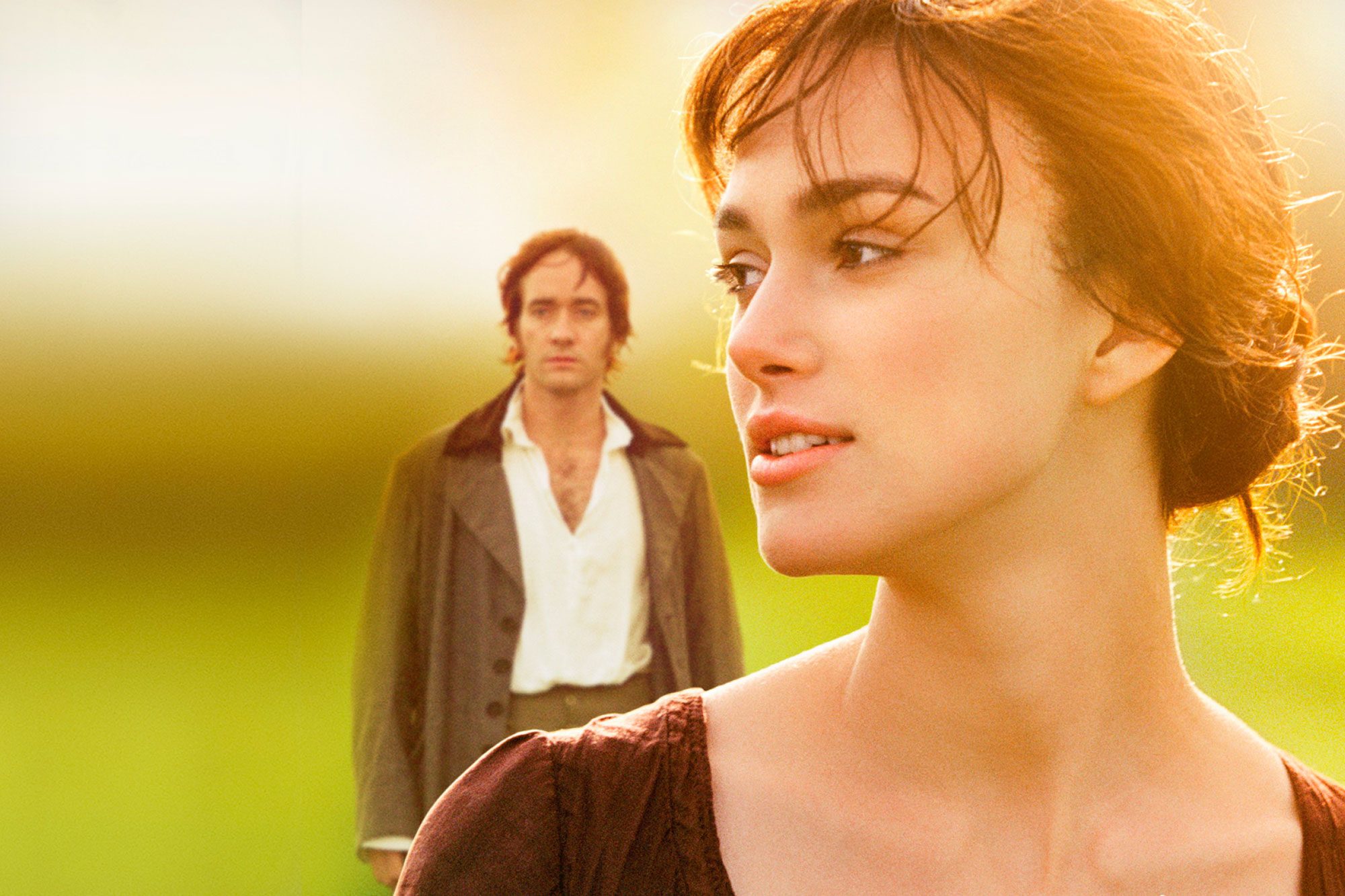 A woman gazes thoughtfully while a man stands in the background, set in a sunlit, blurry field.