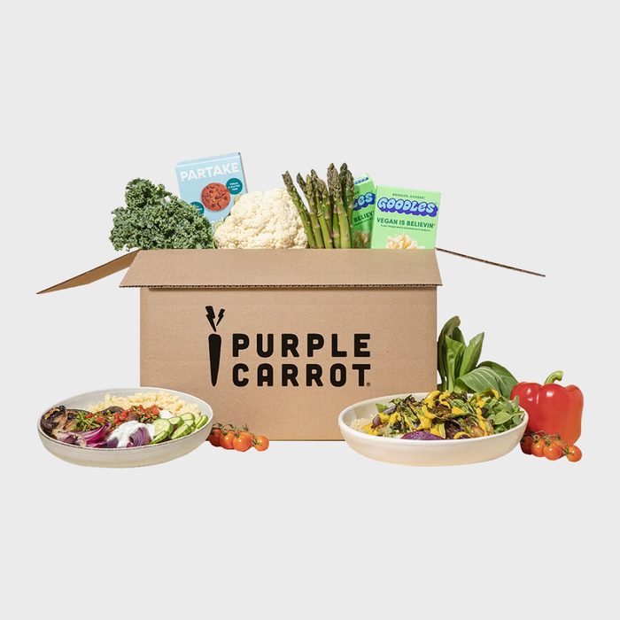 Box holds fresh vegetables and packaged snacks; placed between two plates of colorful salads, surrounded by small tomatoes and a red pepper.