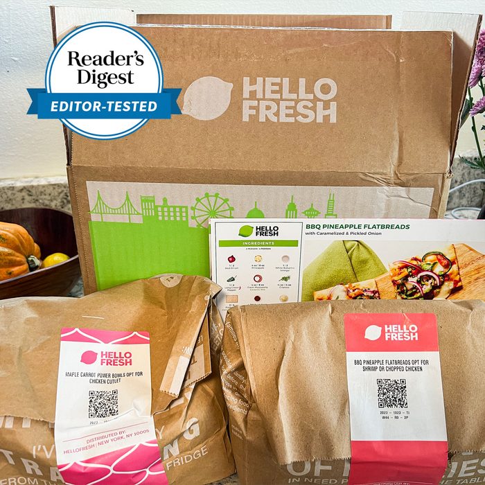 HelloFresh meal kit sits unopened, displaying BBQ Pineapple Flatbreads recipe card and ingredient bags, surrounded by kitchen dÃ©cor.