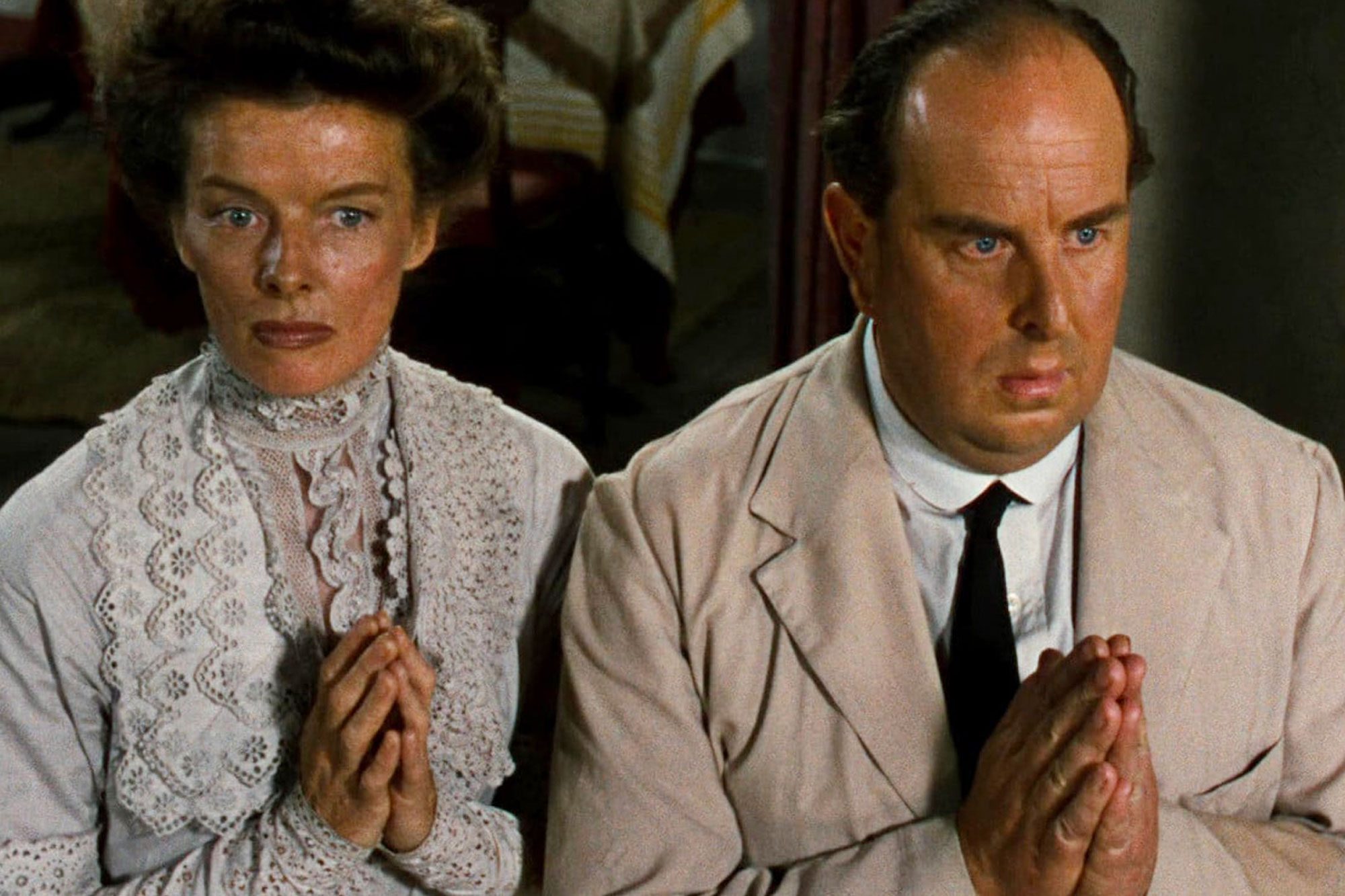 Two people sit with hands in prayer position, dressed in formal attire, inside a dimly lit room.