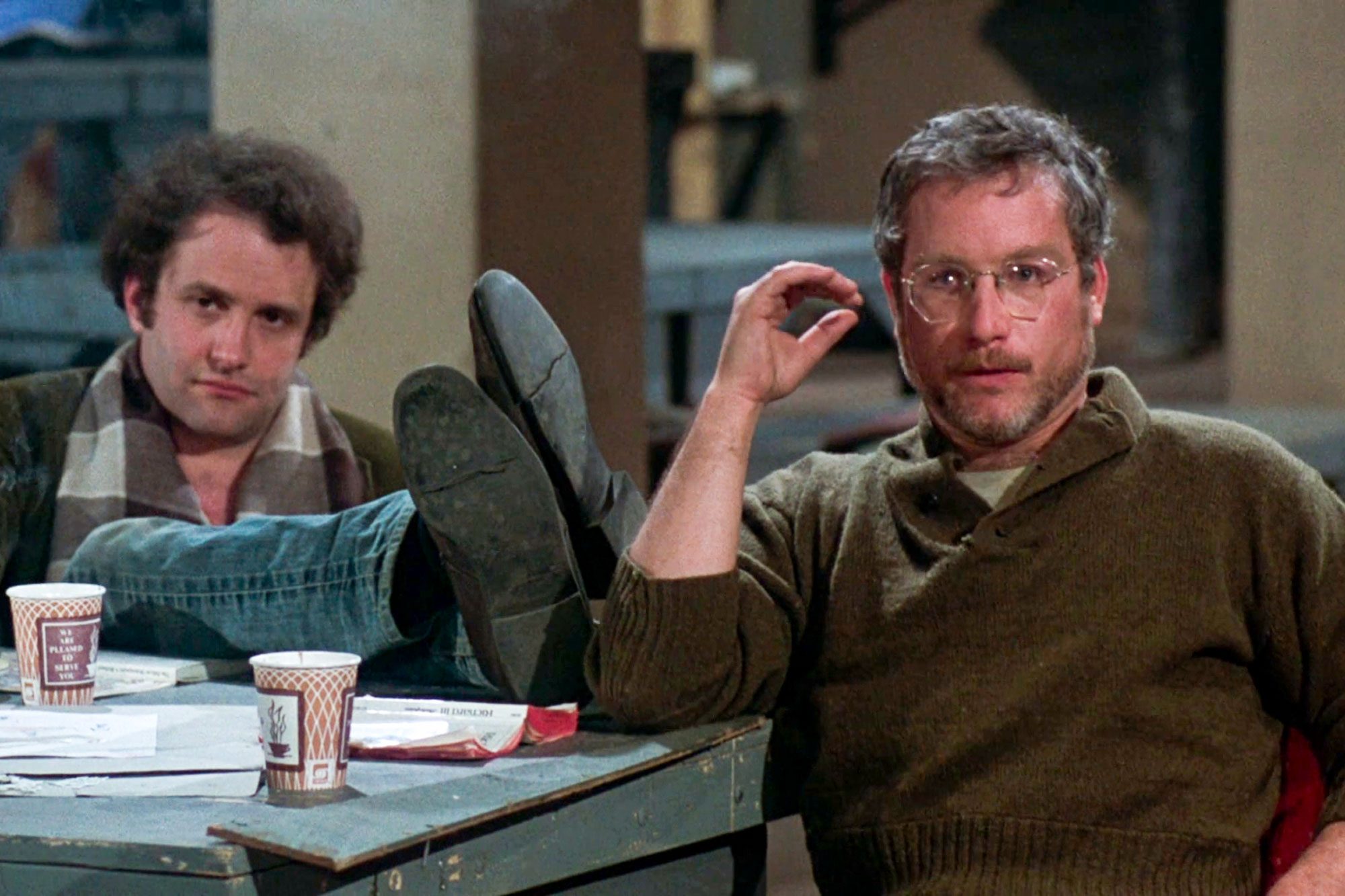 Two people sit relaxed; one with feet on table, next to coffee cups and papers in a casual, indoor setting.