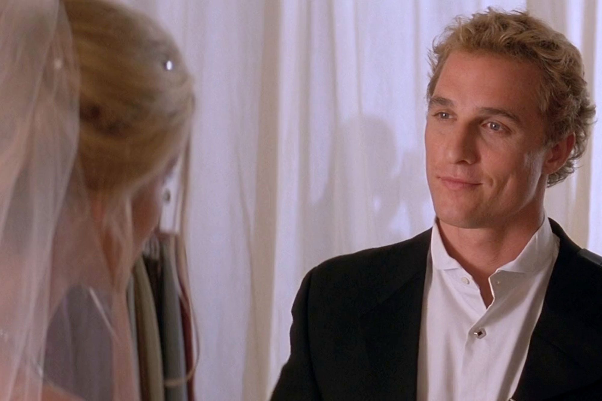 A man in a suit smiles at a woman in wedding attire, set against white hanging drapes.
