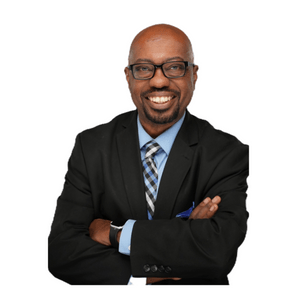 A smiling man crosses his arms, wearing glasses, a black suit, and a blue checkered tie, against a plain white background.