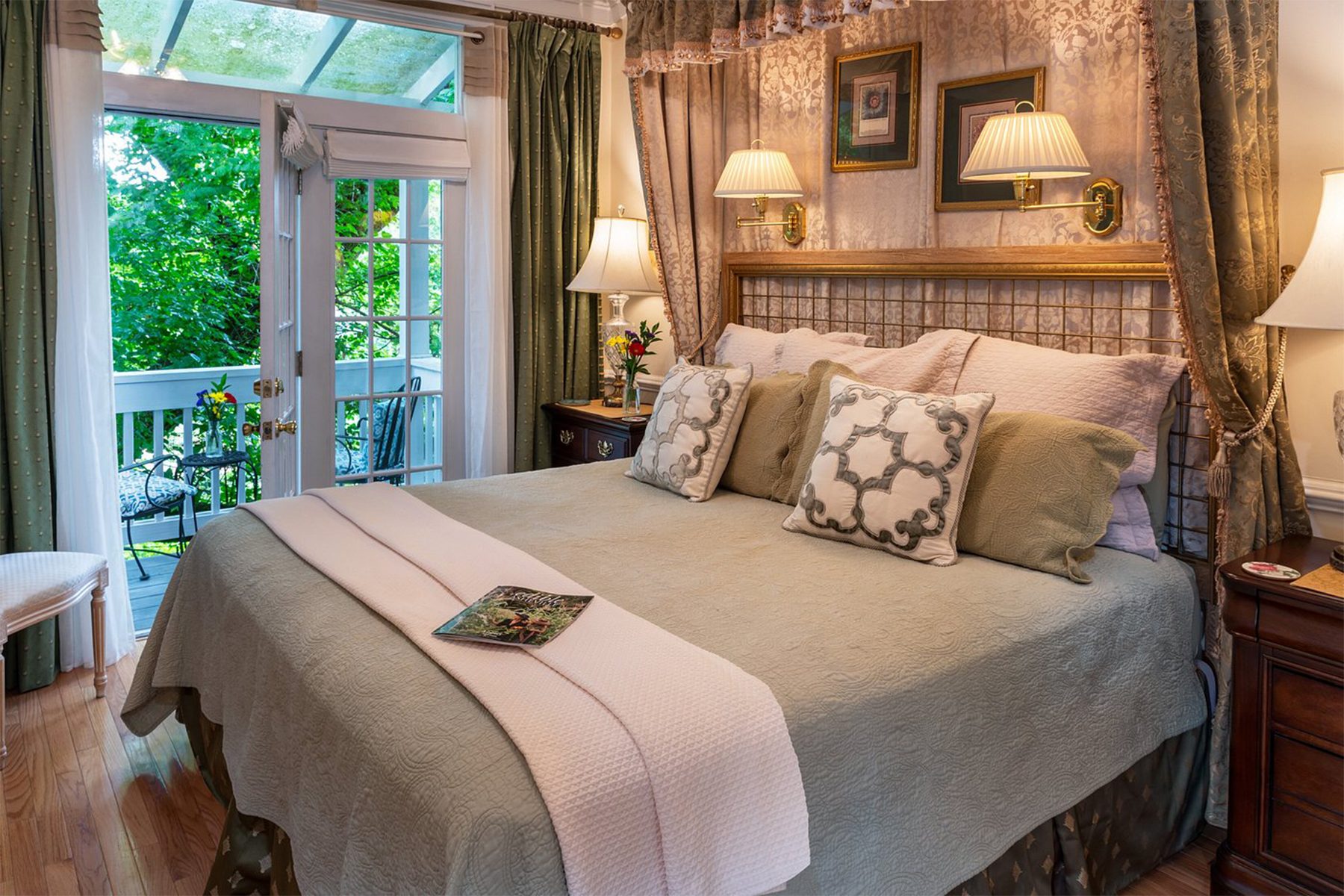 A bed with pillows and a blanket faces French doors, opening to a leafy outdoor area, surrounded by curtains and lamps.