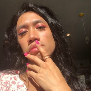 Person with pink makeup and nails touching lips, in soft sunlight, against an indoor background with ceiling lights.