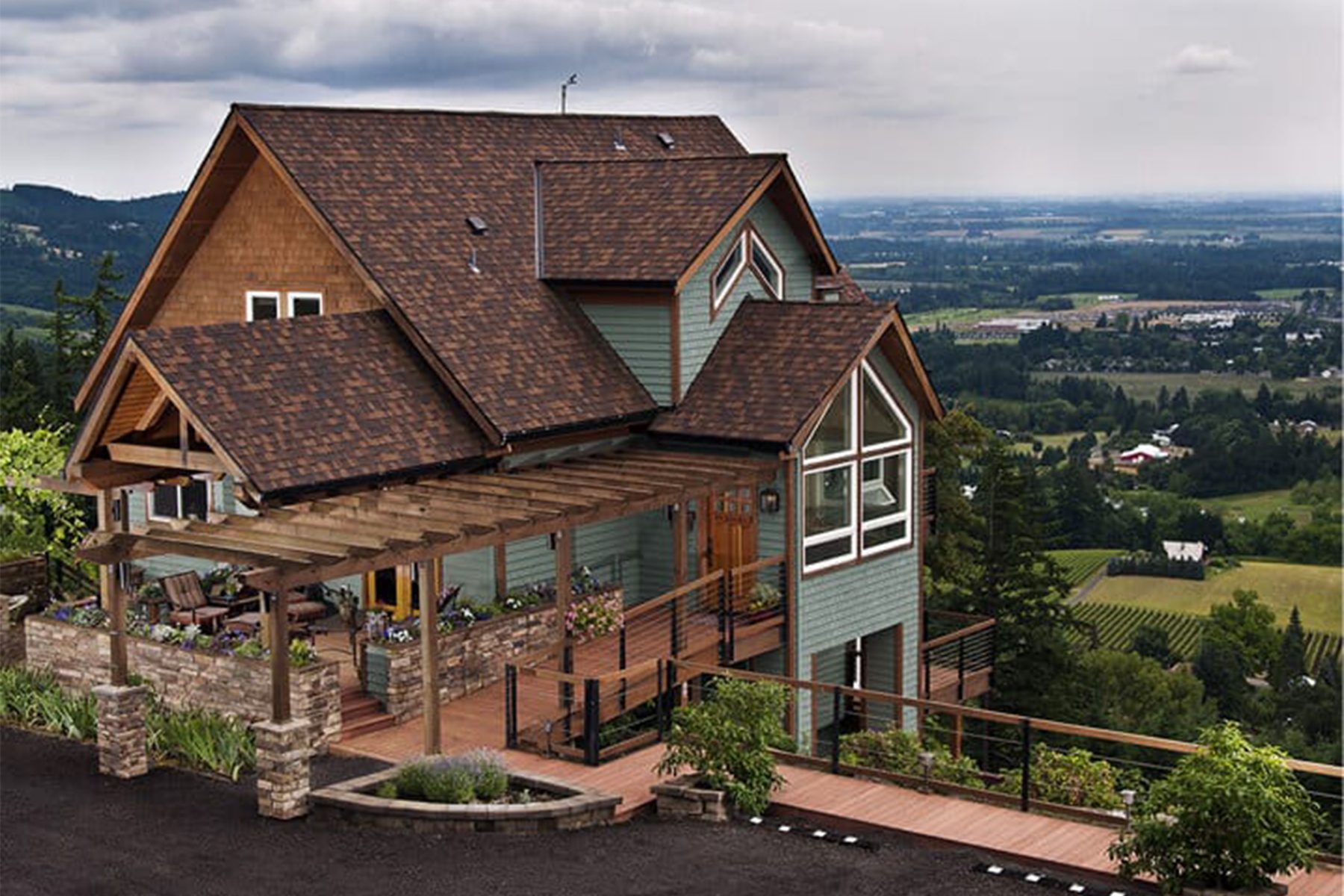 House overlooks a green valley, featuring brown roof and wooden deck with pergola. Surrounding landscape includes hills and distant buildings under a cloudy sky.