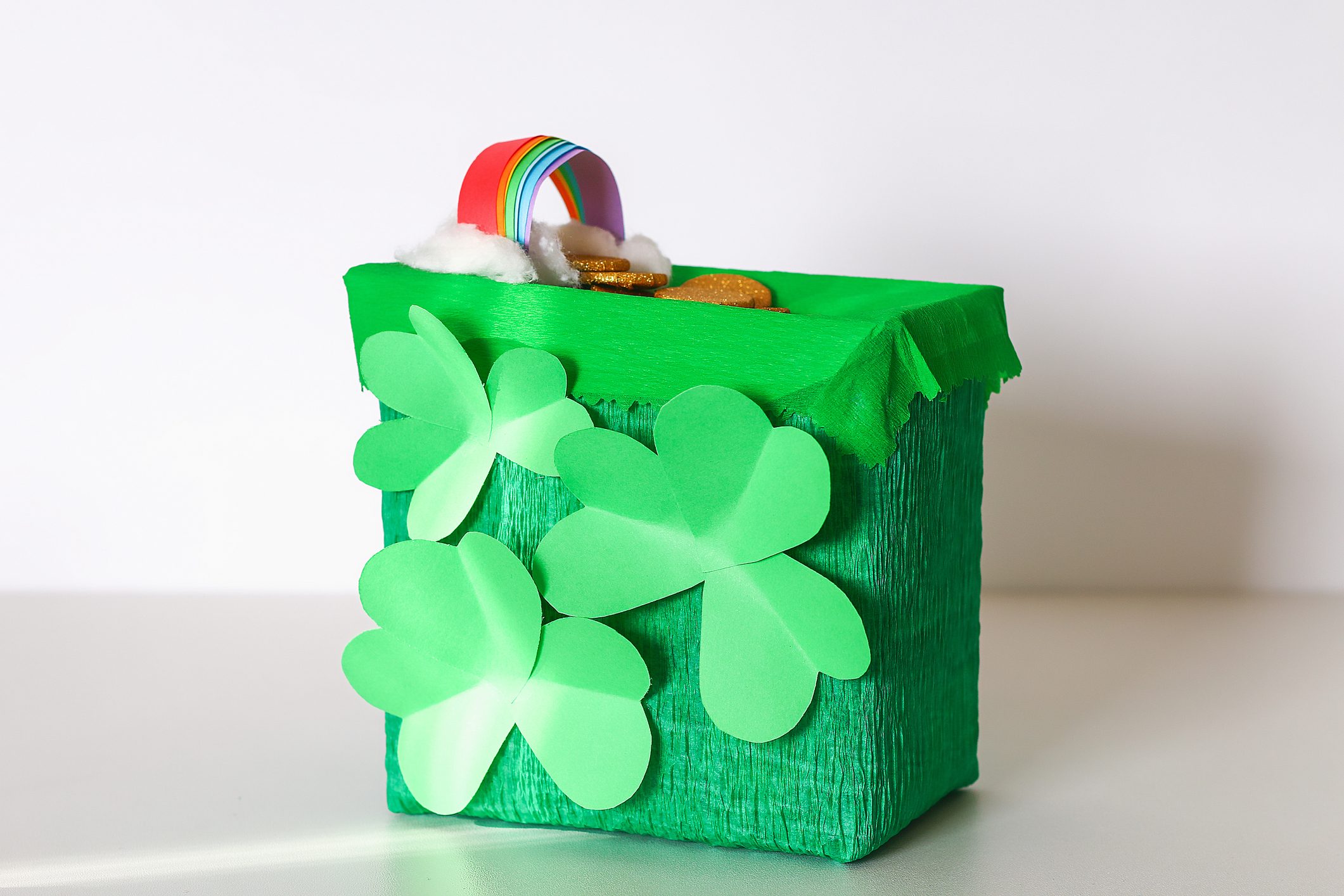 Green box displays paper shamrocks, topped with a rainbow arch and faux gold coins, set on a plain background.