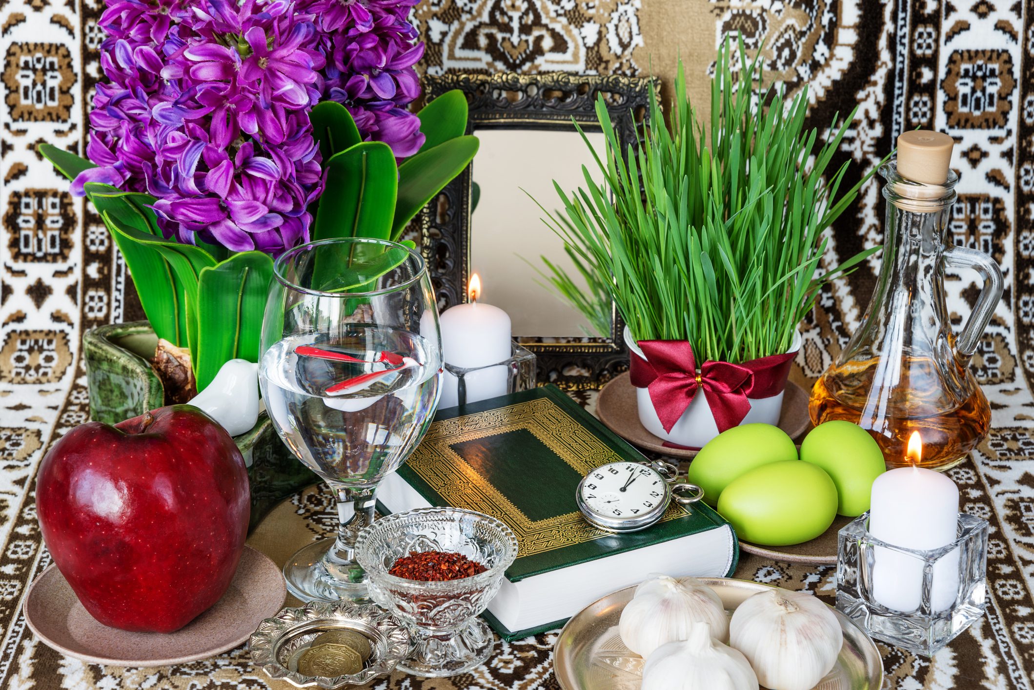 Objects arranged on a patterned tablecloth: apple sits beside hyacinth, water glass with fish, green wheatgrass, candles, garlic, eggs, book, and coins.