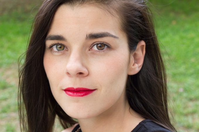 Portrait of woman with heterochromia iridum eyes brown and green and red lipstick looking to the camera confident and relaxed on an outdoors grass background.