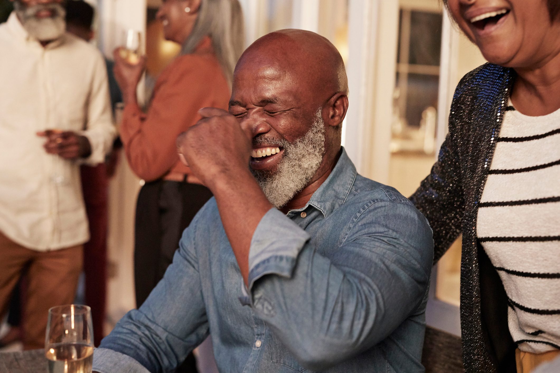 Man laughing heartily, surrounded by people in a social gathering, some holding drinks, indoors.