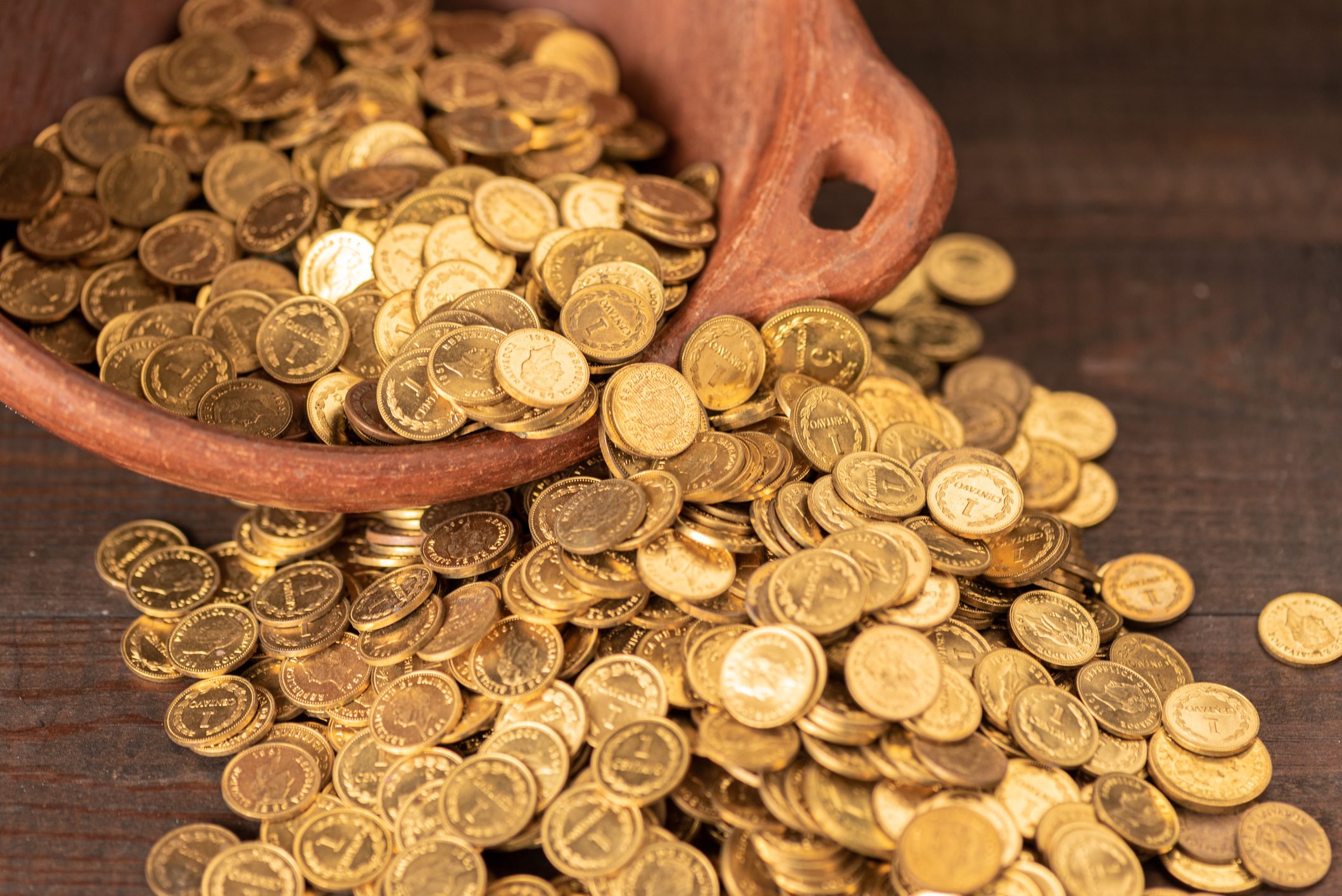 Coins spill from a clay pot, creating a scattered pile on a wooden surface.