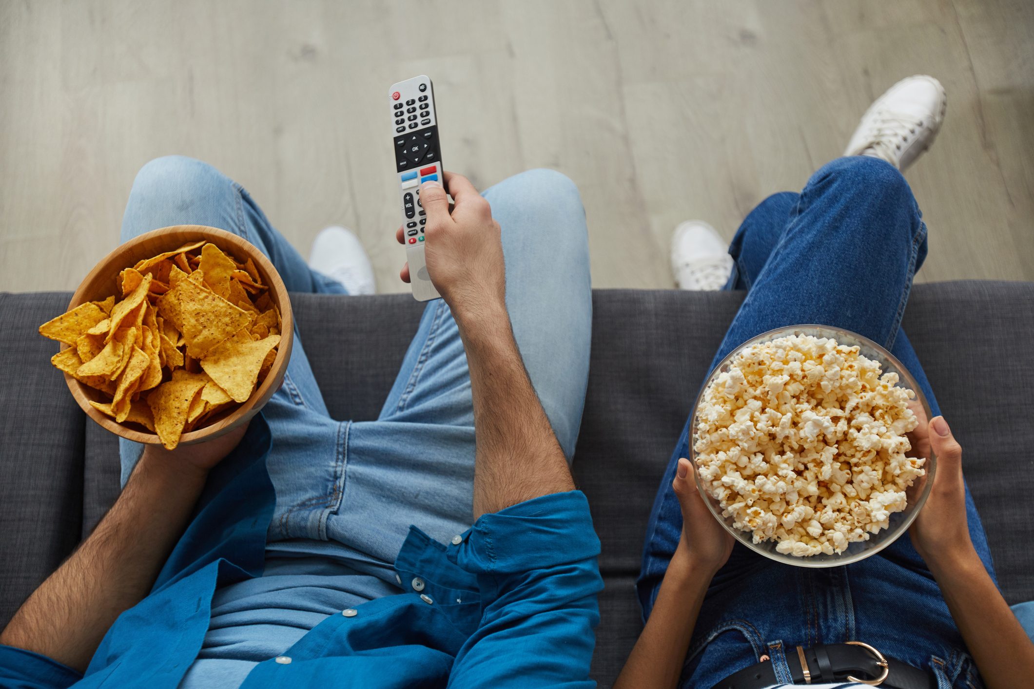 People sit on a couch holding snacks and a remote, enjoying chips and popcorn in a relaxed living room setting.