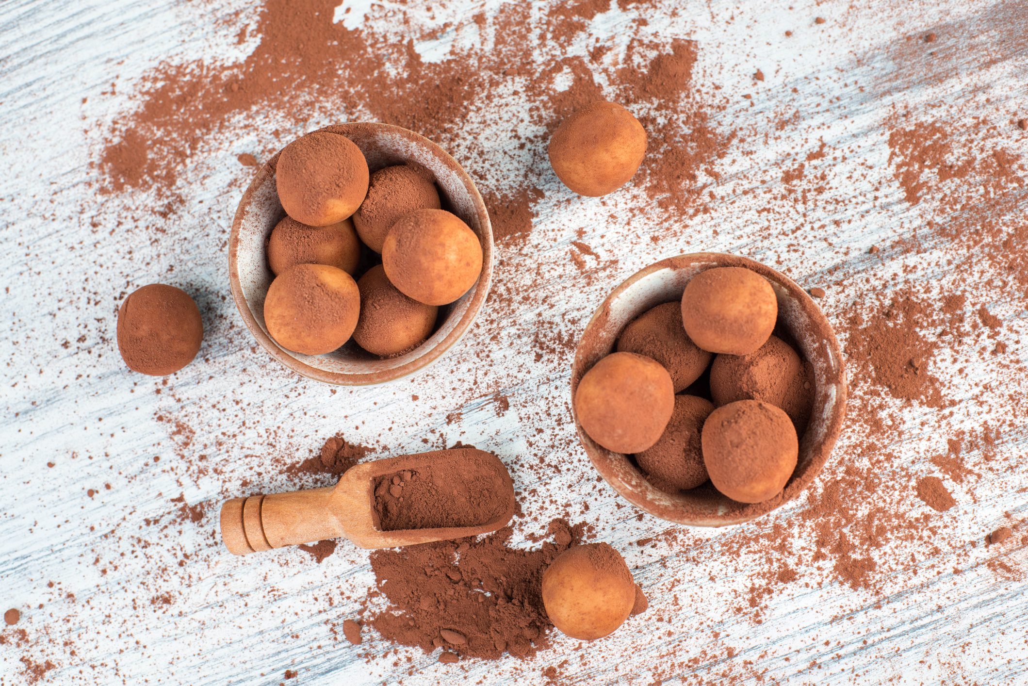 Cocoa-dusted truffles rest in and around bowls on a powdered wooden surface, accompanied by a wooden scoop holding more cocoa powder.