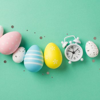 Colorful Easter eggs are arranged in a line with a small clock on a mint green background, surrounded by scattered confetti.