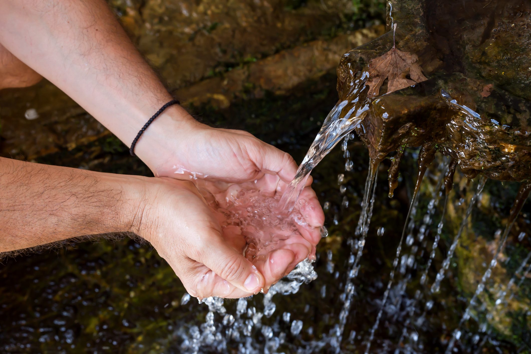 Hands cupping clear water flowing from a mossy stone fountain outdoors.