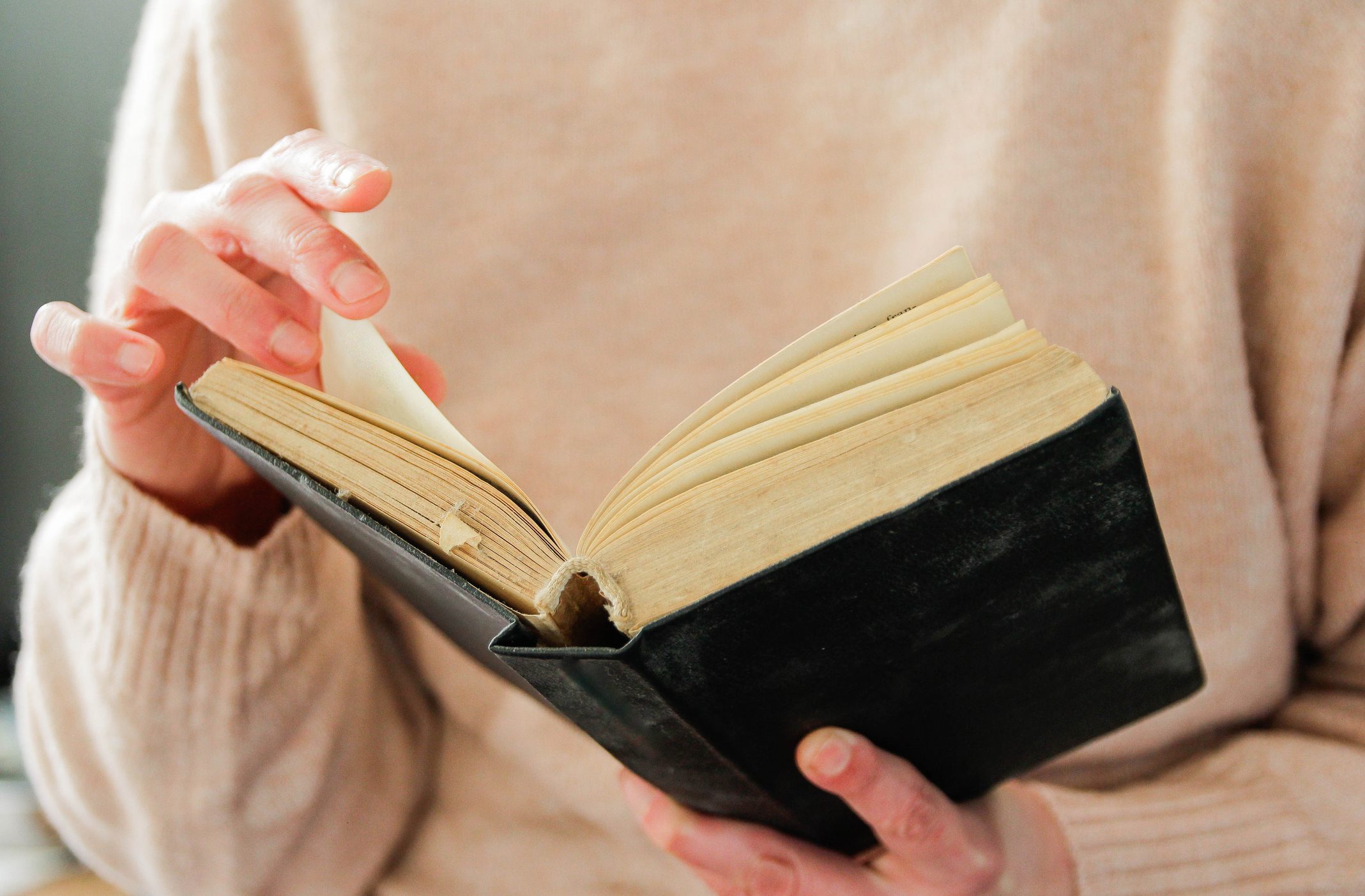 Hands turning pages of an old book, against a blurred beige sweater background.