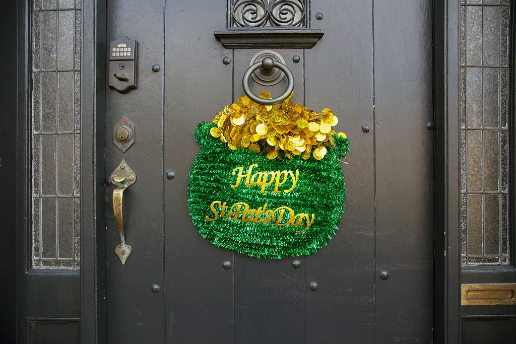 Decorative wreath hangs with gold coins on a black door; text reads "Happy St. Patrickâ€™s Day." Surroundings feature a keypad and brass handle.