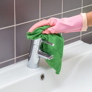 A gloved hand cleans a shiny faucet with a green cloth, set against gray tiled wall in a bathroom.