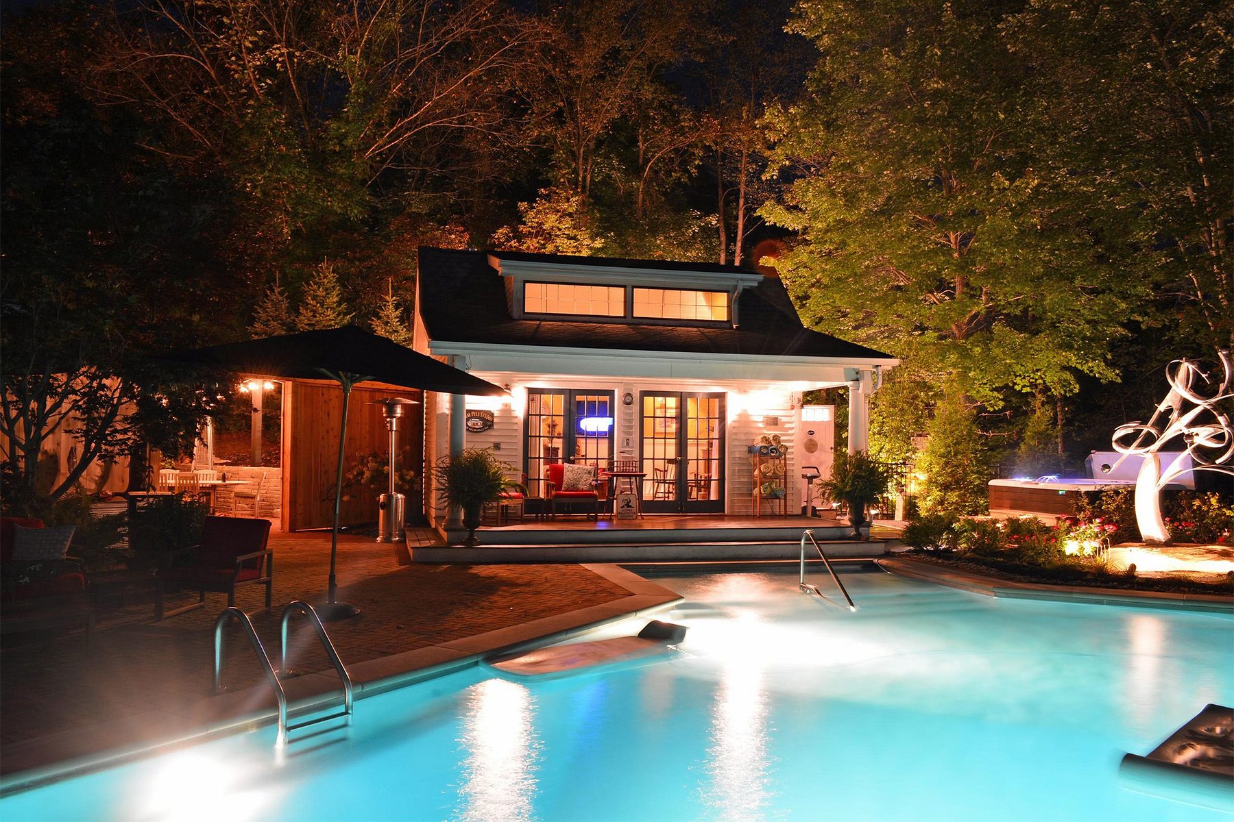 Pool glows softly, beside a lit pool house with patio furniture, surrounded by trees and illuminated sculptures in the night garden setting.