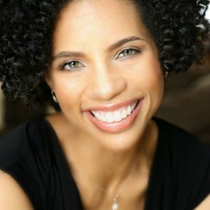 Smiling person with curly hair looks directly at the camera, wearing a black top, set against a neutral indoor background.