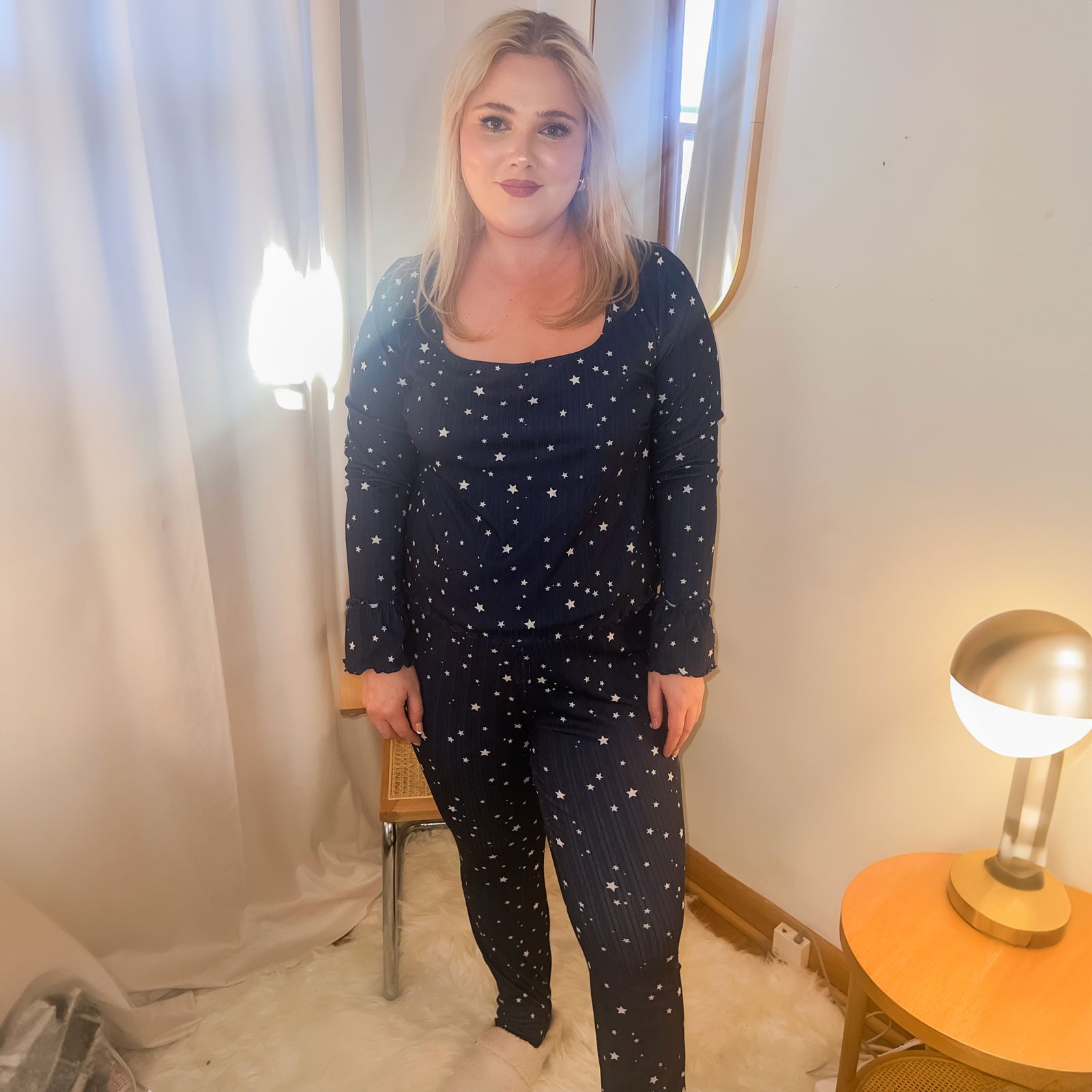 A person stands, wearing star-patterned pajamas, in a cozy room with a glowing lamp on a small wooden table, and light-filtering curtains.