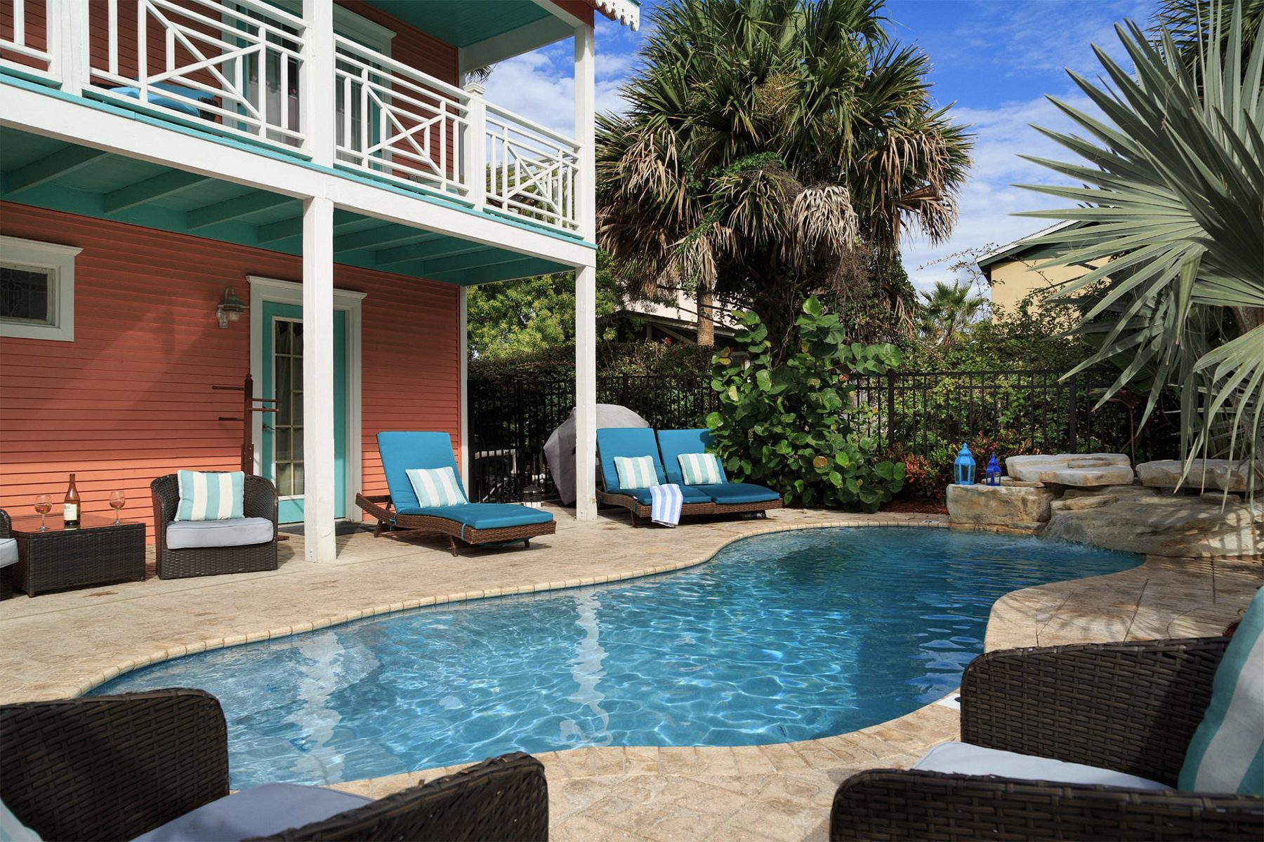 A swimming pool glistens under the sun, surrounded by blue loungers and palm trees, adjacent to a coral-painted house with a balcony.