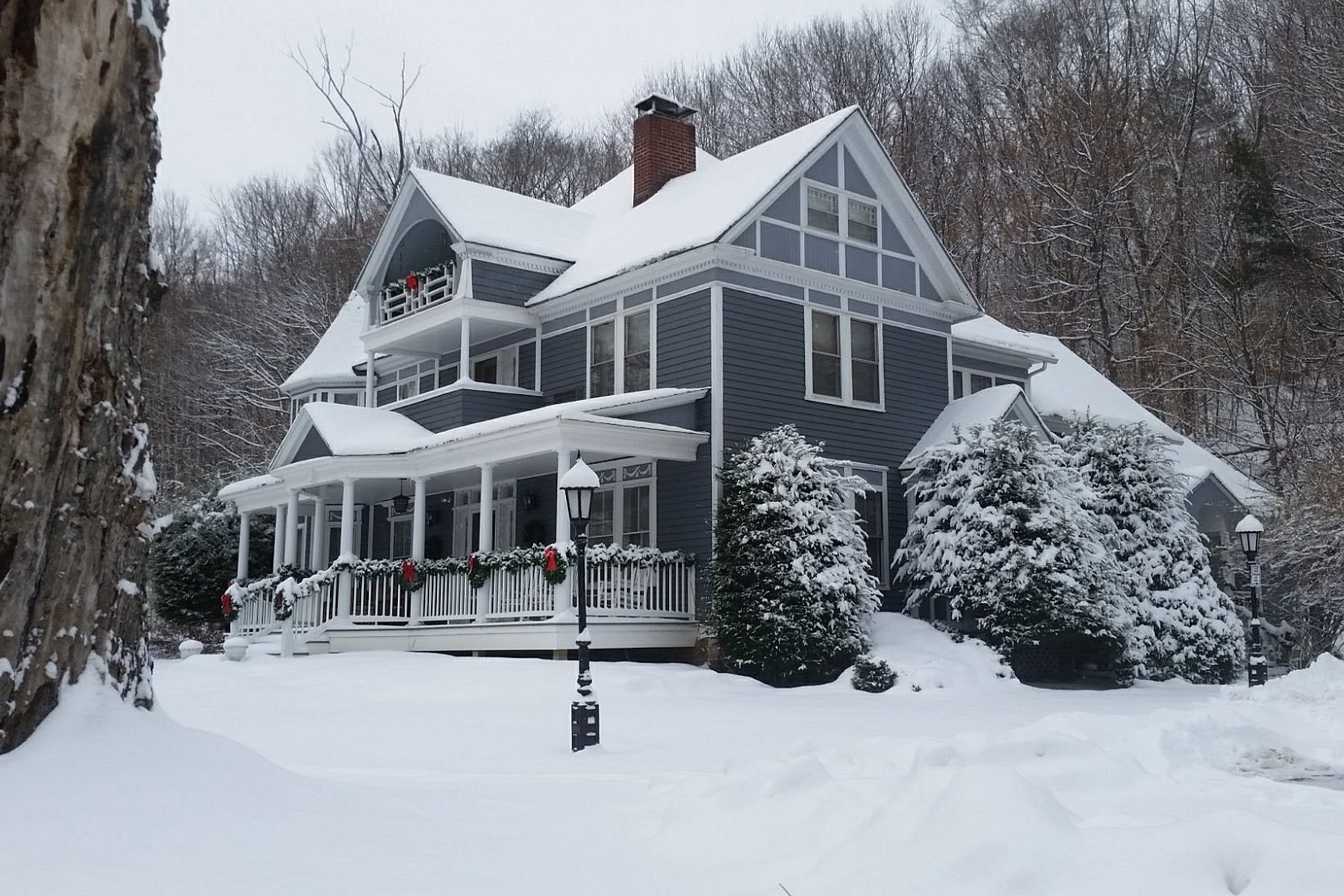 House covered in snow; festive garlands decorate the porch railing amidst wintry trees and a snow-blanketed yard.