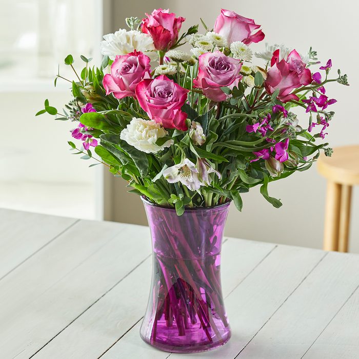 Pink and white flowers are arranged in a purple vase, sitting on a wooden table in a bright room.