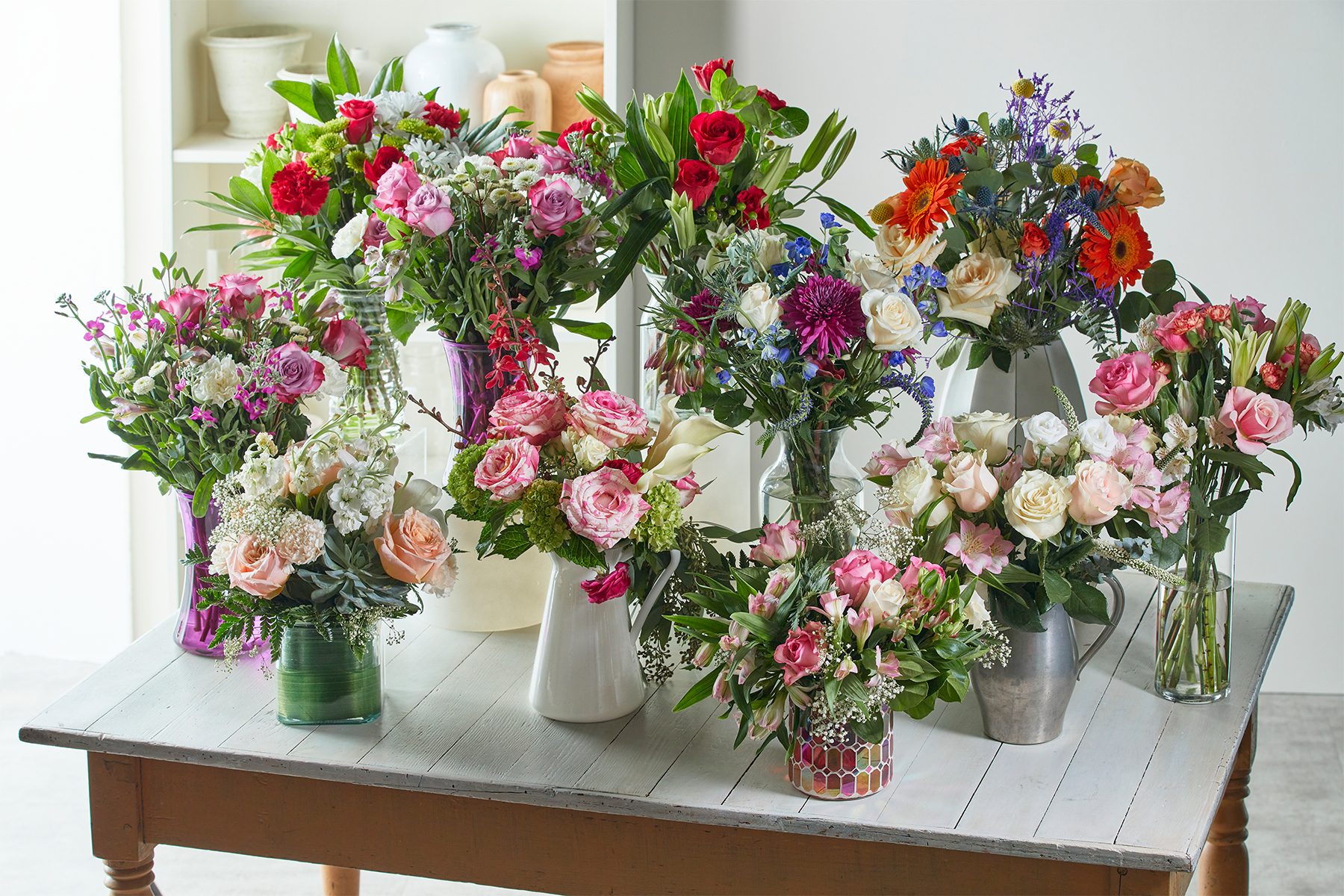 Flower arrangements in colorful vases stand on a wooden table, surrounded by a bright room with pottery on shelves.