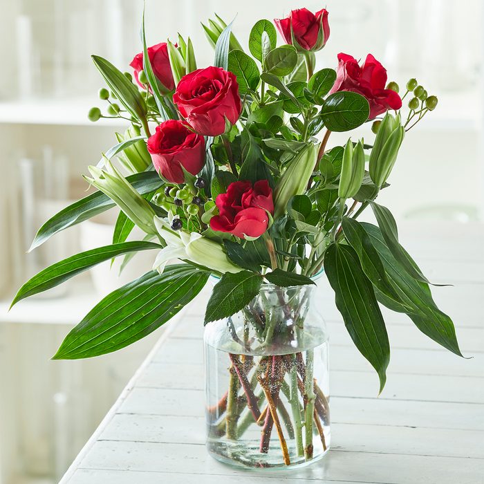 A glass vase holds red roses and green foliage, resting on a white wooden table in a bright room.