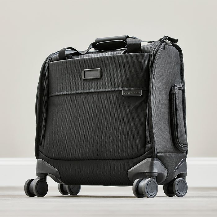 A black suitcase stands on four wheels in a minimalistic indoor setting. A label reads 