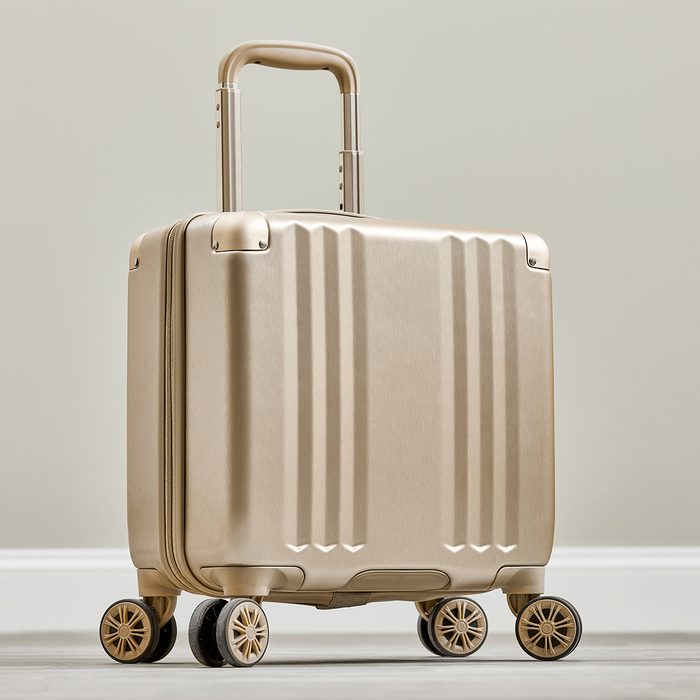 A beige suitcase stands upright with its handle extended, positioned on four wheels, against a simple indoor background.