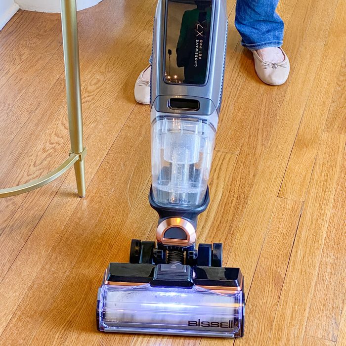 Vacuum cleaner moves across wooden floor; person wearing jeans and beige shoes operates it near a metal table leg.